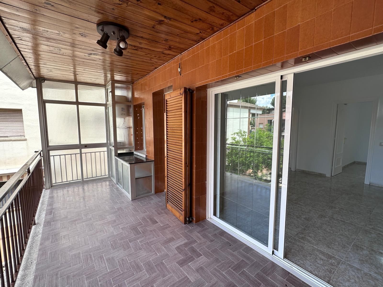 Balcón con piso de ladrillo, contraventanas marrones y puerta corrediza de vidrio a una habitación vacía.