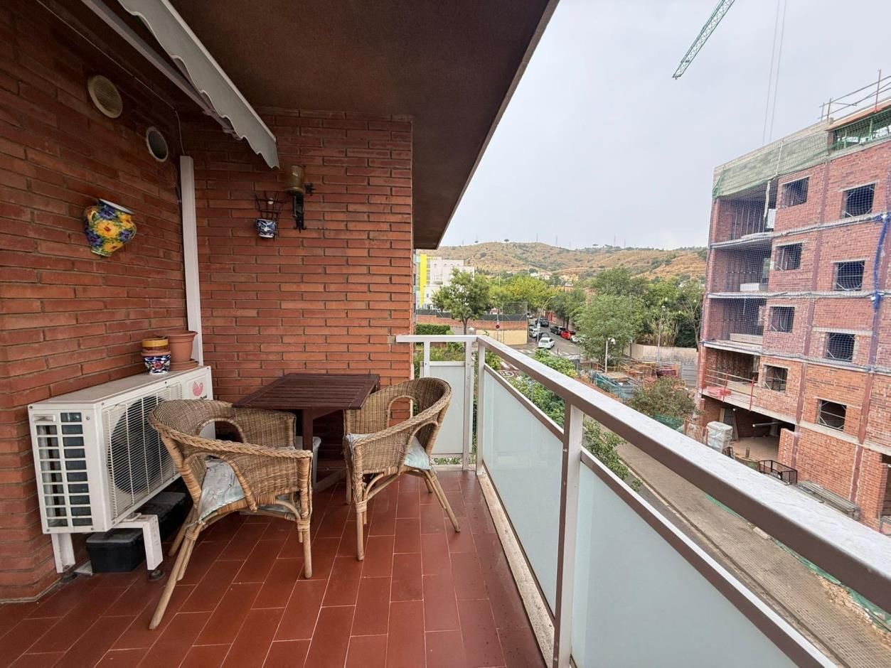 Balcón con mesa y sillas, con vistas a la construcción del edificio y a una colina. Pared de ladrillo rojo y aire acondicionado visibles.