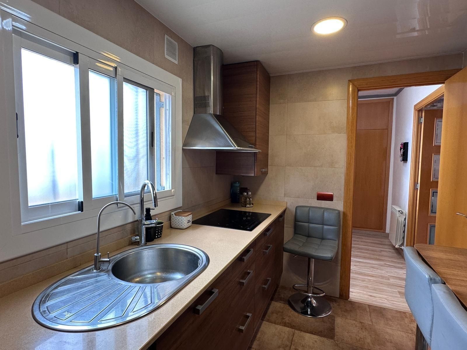 Cocina con fregadero, estufa y extractor de acero inoxidable. Gabinetes de madera, paredes color canela y un pequeño taburete.