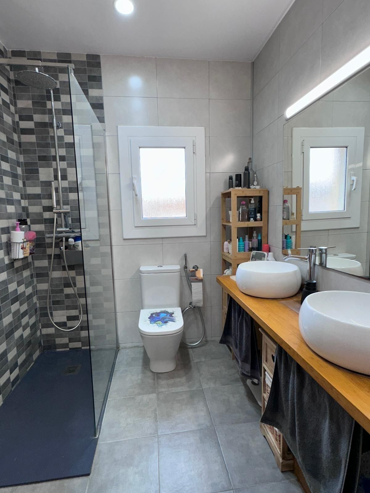 Baño con ducha, inodoro y lavabo doble. Azulejos grises y blancos, encimera de madera y tonos neutros.