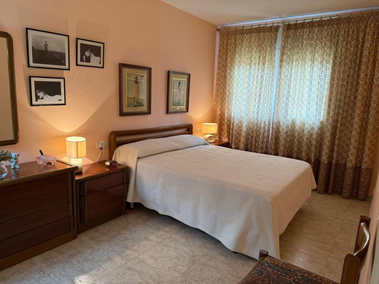 Dormitorio con paredes color melocotón, cama y cortinas con estampado floral. Hay una cómoda, mesitas de noche y cuadros.