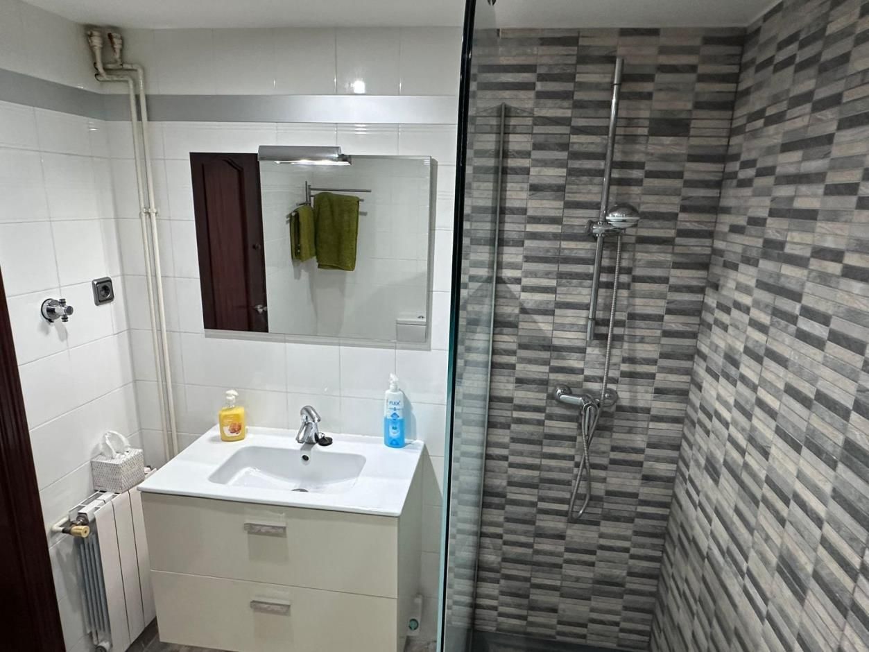 Pequeño baño con tocador blanco, cabina de ducha y paredes de azulejos grises.