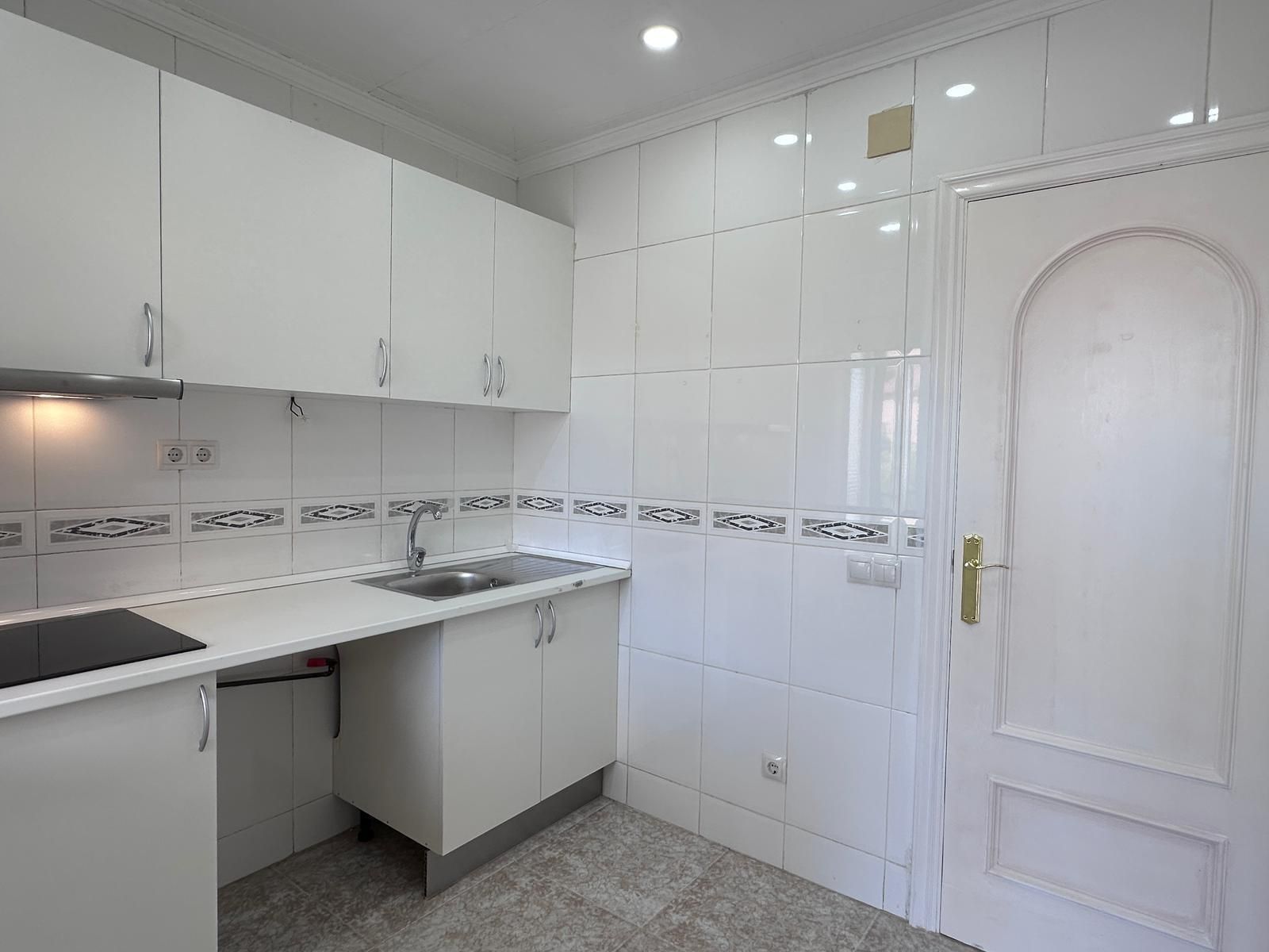 Cocina blanca con gabinetes, fregadero y puerta, paredes de azulejos y piso gris.
