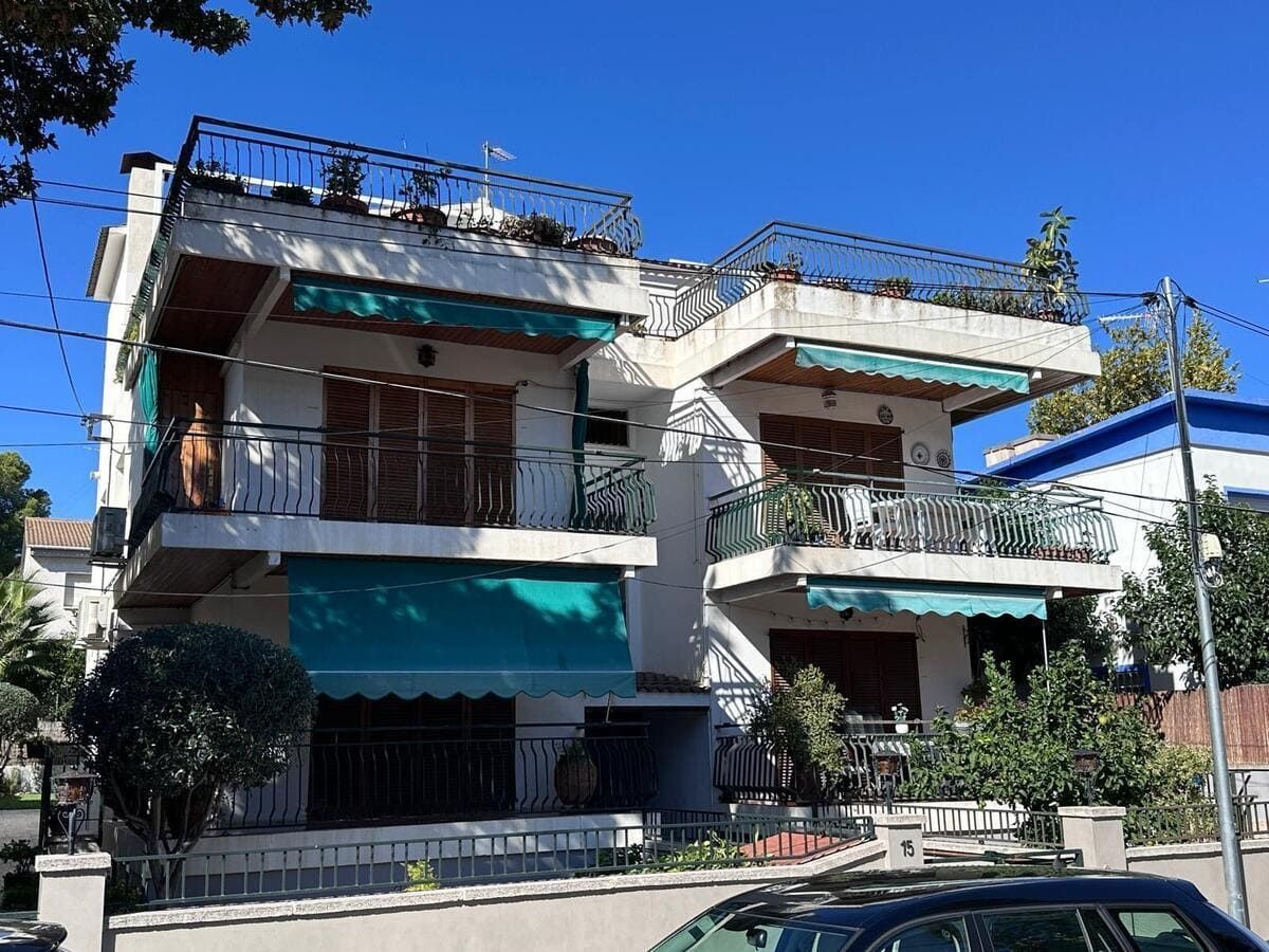 Edificio blanco de dos pisos con balcones, toldos y jardineras en la azotea bajo un cielo azul claro.