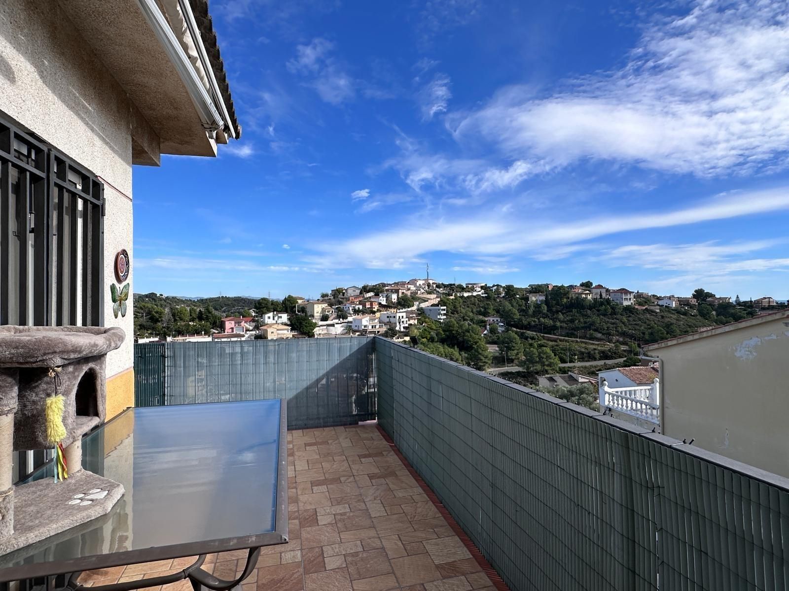 Terraza con vistas a un pueblo en la ladera bajo un cielo azul radiante. Se ven una valla verde, una mesa y un árbol para gatos.