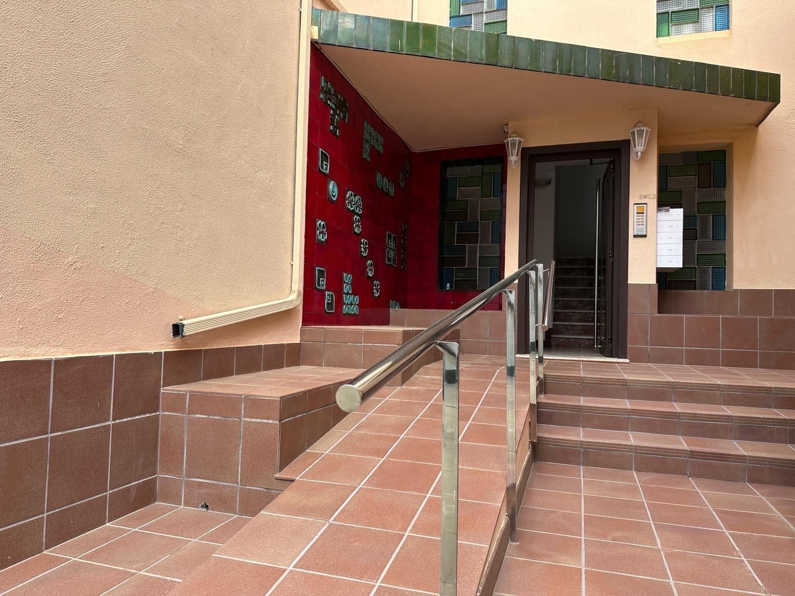 Entrada con rampa y escaleras, presenta una pared roja, azulejos marrones y un toldo verde.