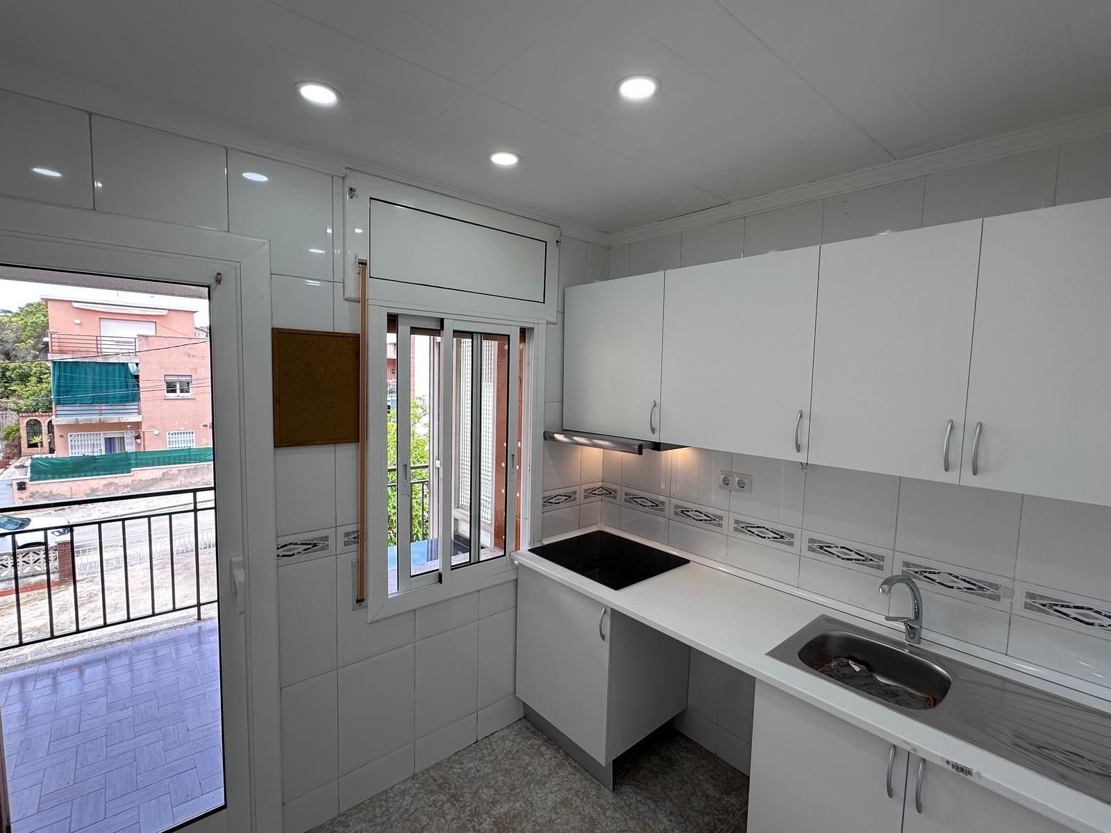 Pequeña cocina blanca con puerta abierta al balcón. Incluye armarios, estufa, fregadero y vista a la calle.