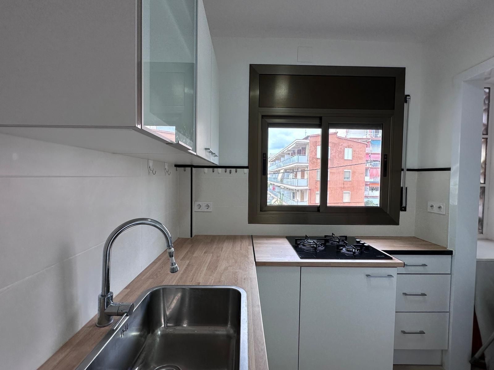 Cocina pequeña y blanca con encimera de madera, fregadero de acero inoxidable y una ventana con vistas a los edificios.