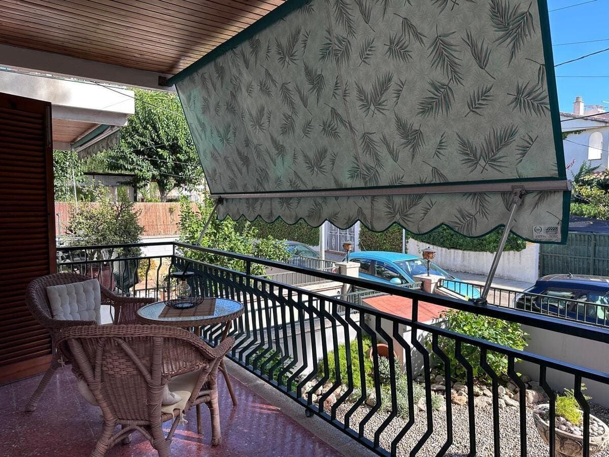 Balcón con toldo, mesa, sillas y barandilla negra. Follaje y coches aparcados al fondo.