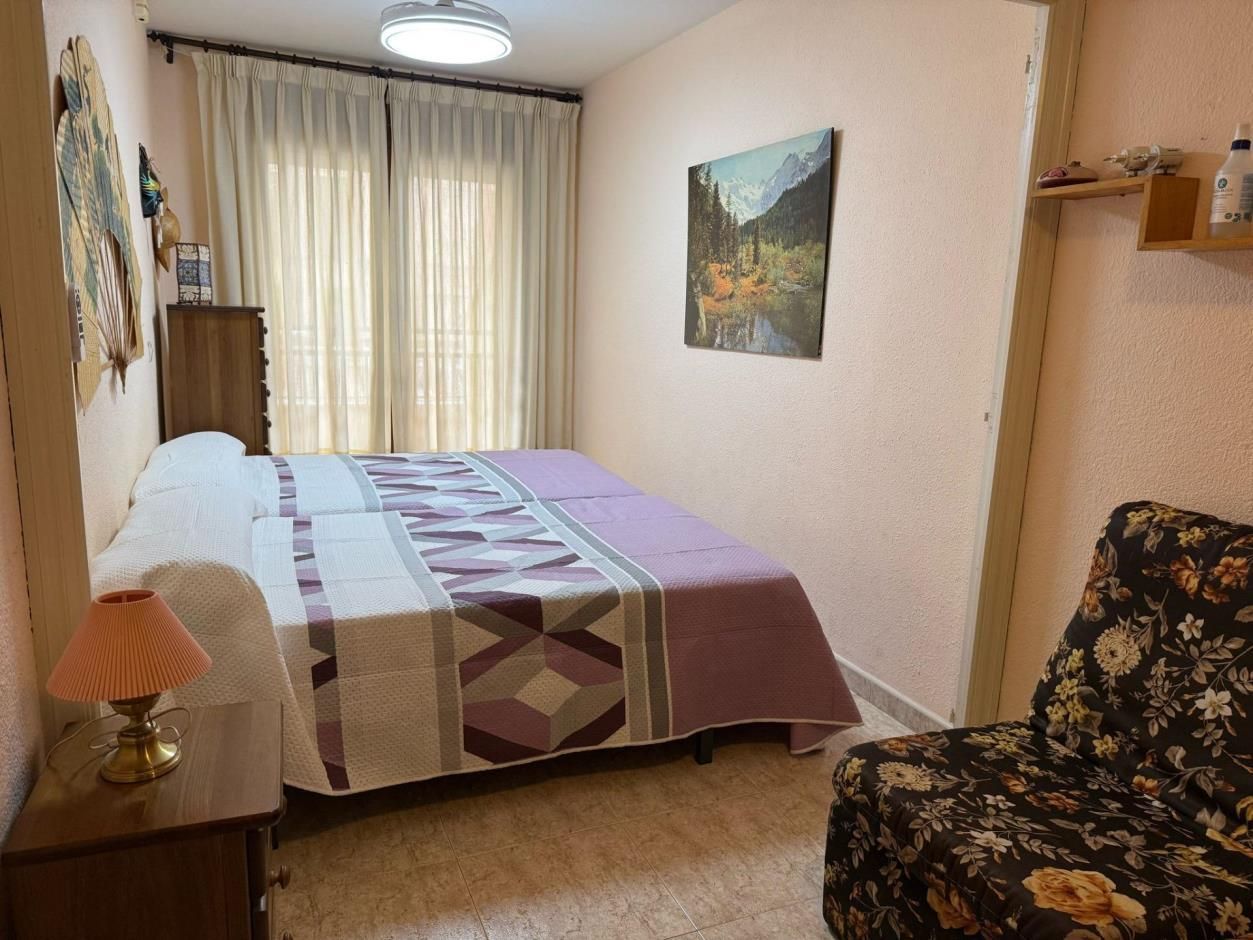 Dormitorio con cama, sofá floral, cómoda de madera y cuadro sobre pared rosa claro.