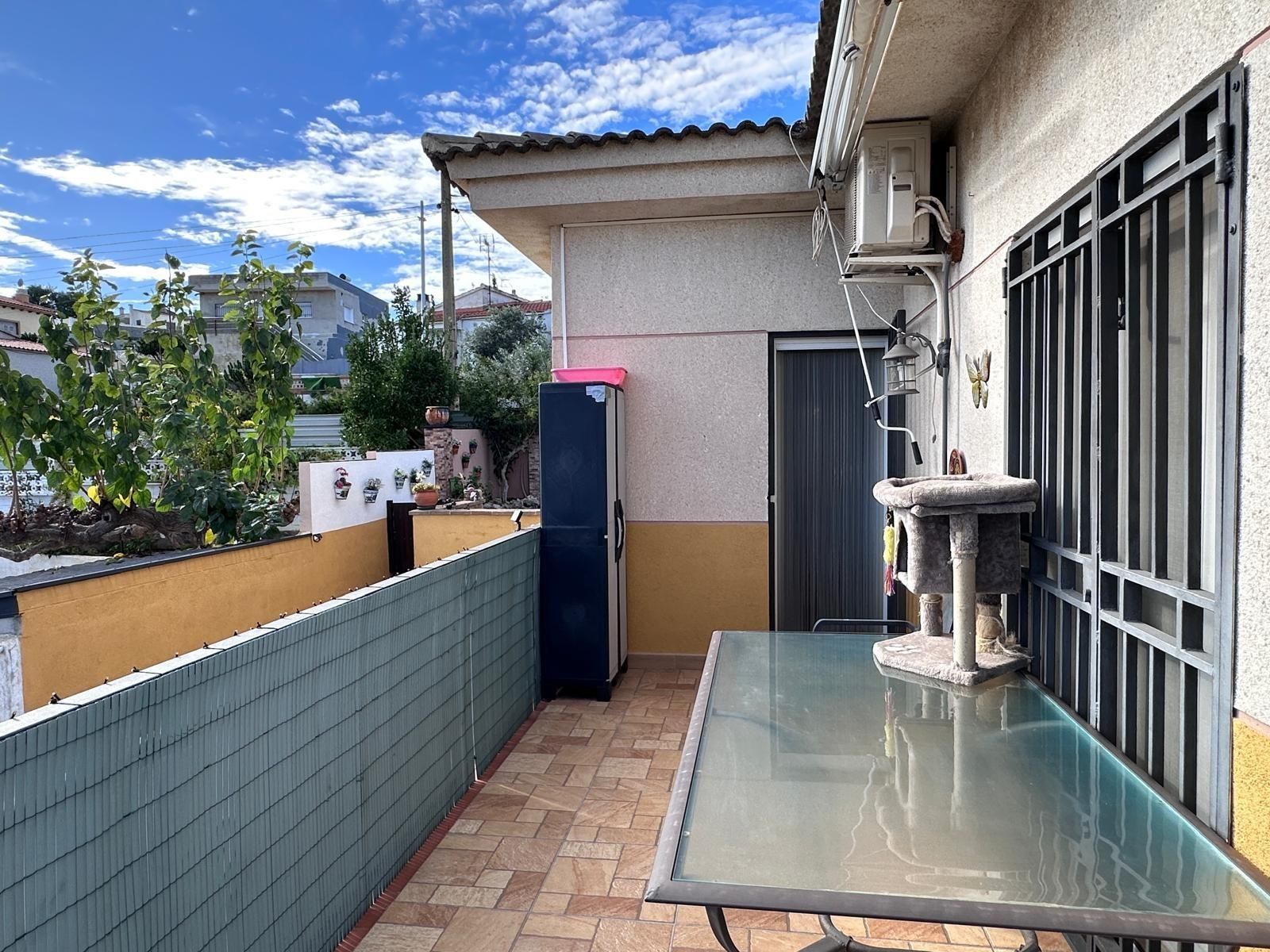 Balcón con mesa de cristal, árbol para gatos y barandilla metálica. Cielo azul con nubes.