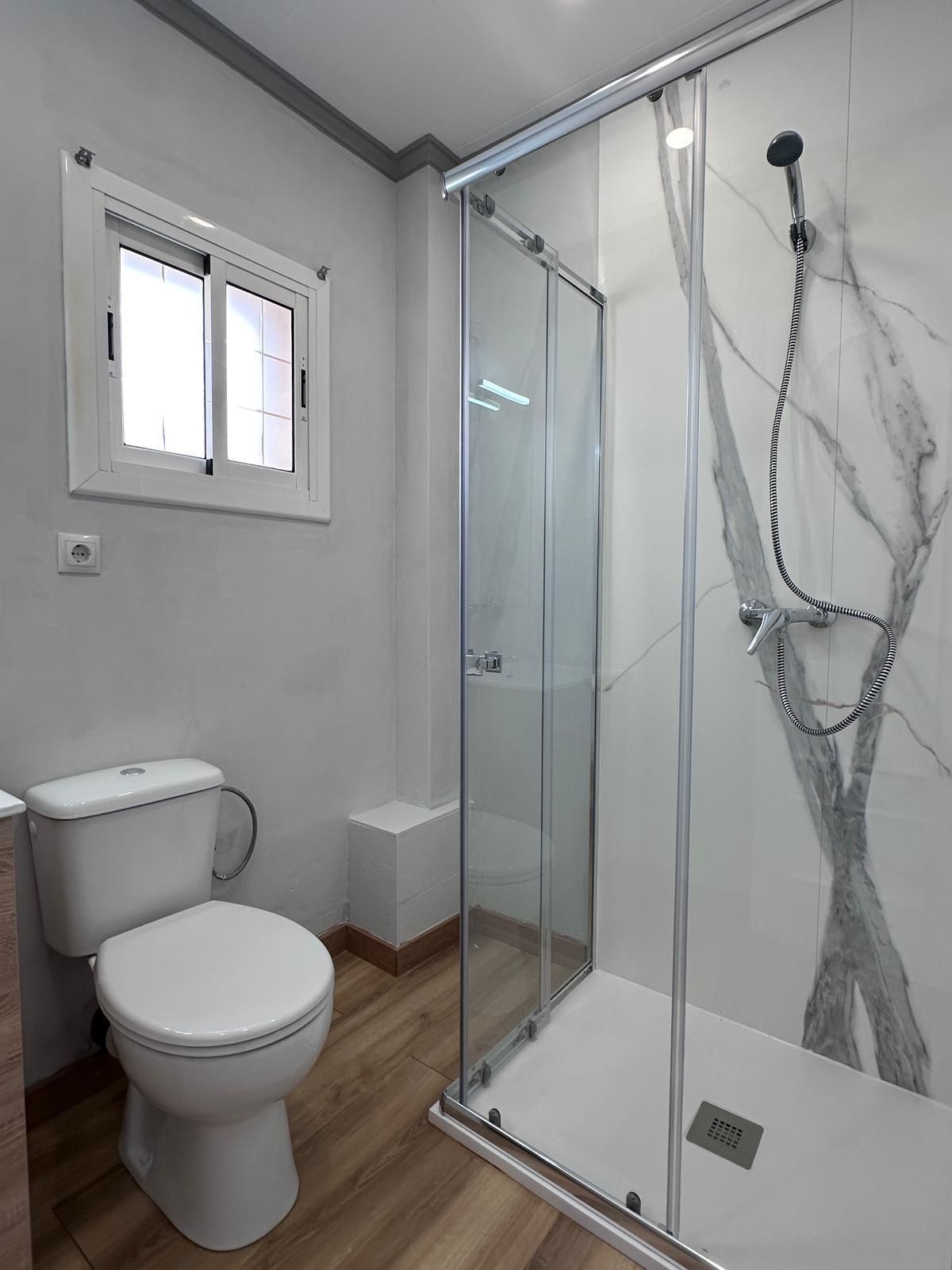 Baño con inodoro, ducha, ventana y diseño de pared símil mármol; paredes gris claro y suelo de madera.