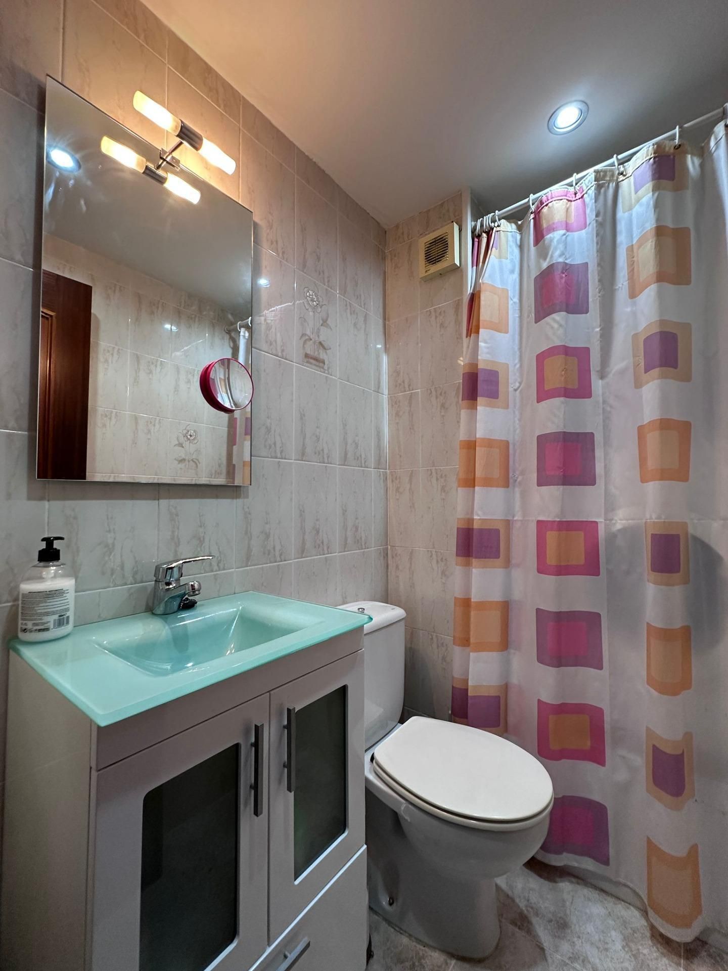 Pequeño baño con tocador blanco, lavabo turquesa, inodoro y cortina de ducha estampada.