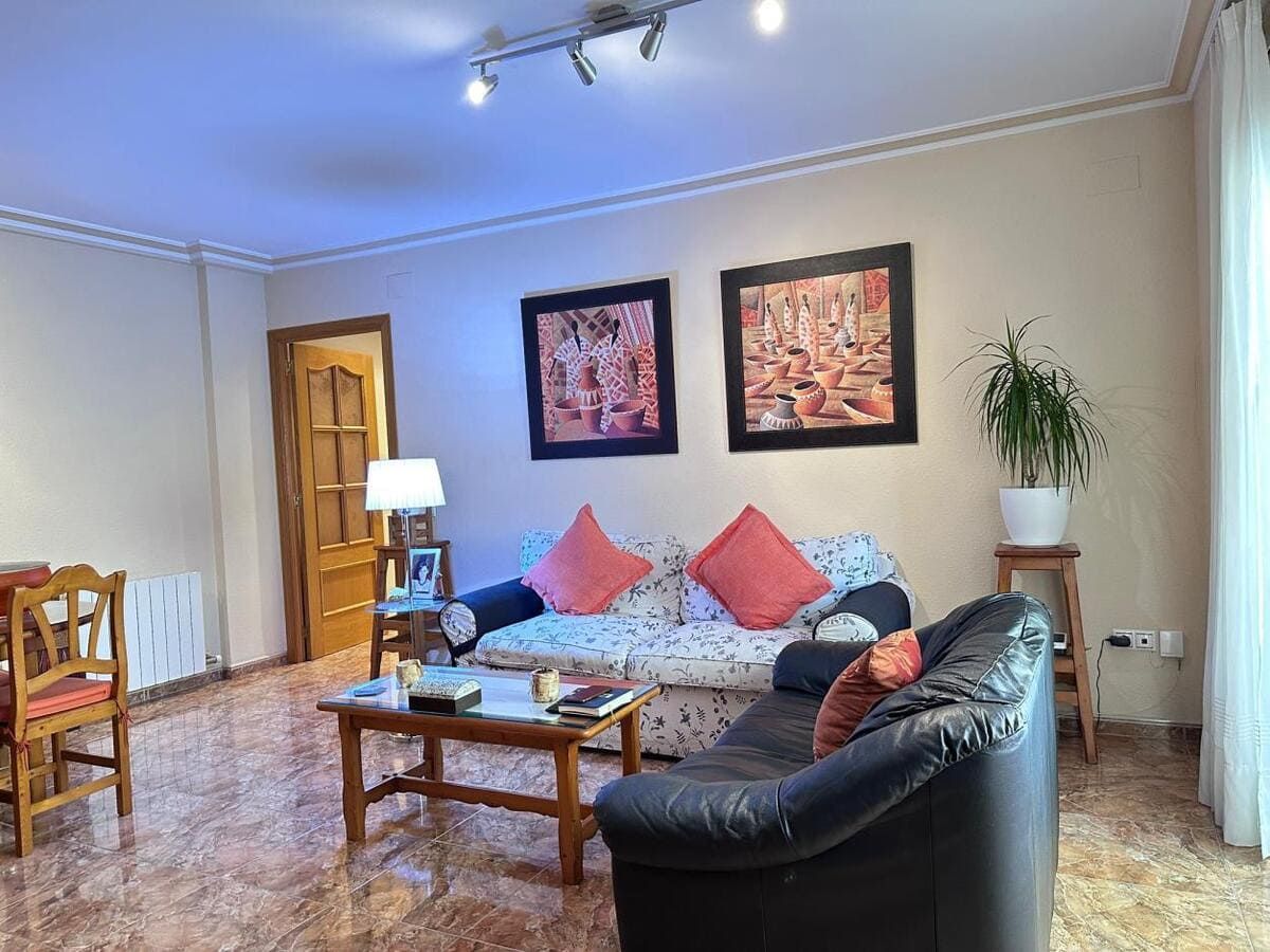 Sala de estar con sofá floral, sofá de dos plazas negro, arte y una planta, con una mesa y sillas a la izquierda.