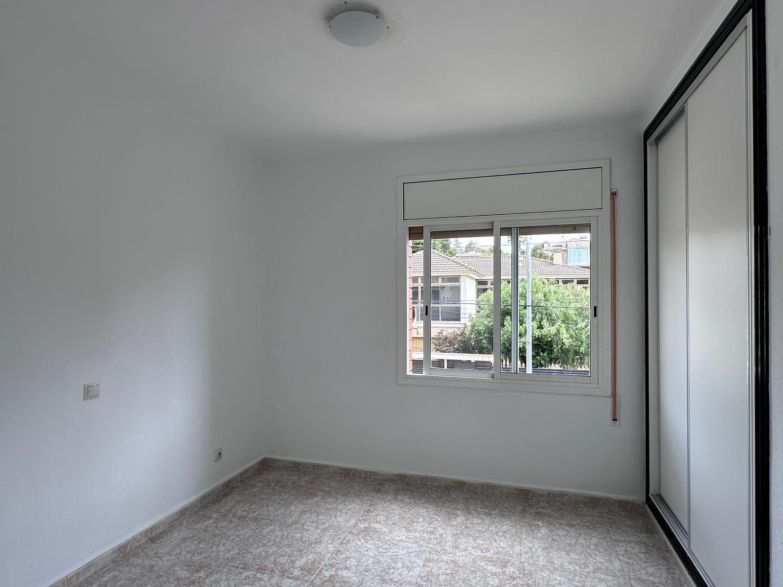 Habitación blanca vacía con ventana y armario, suelo moteado de color marrón.