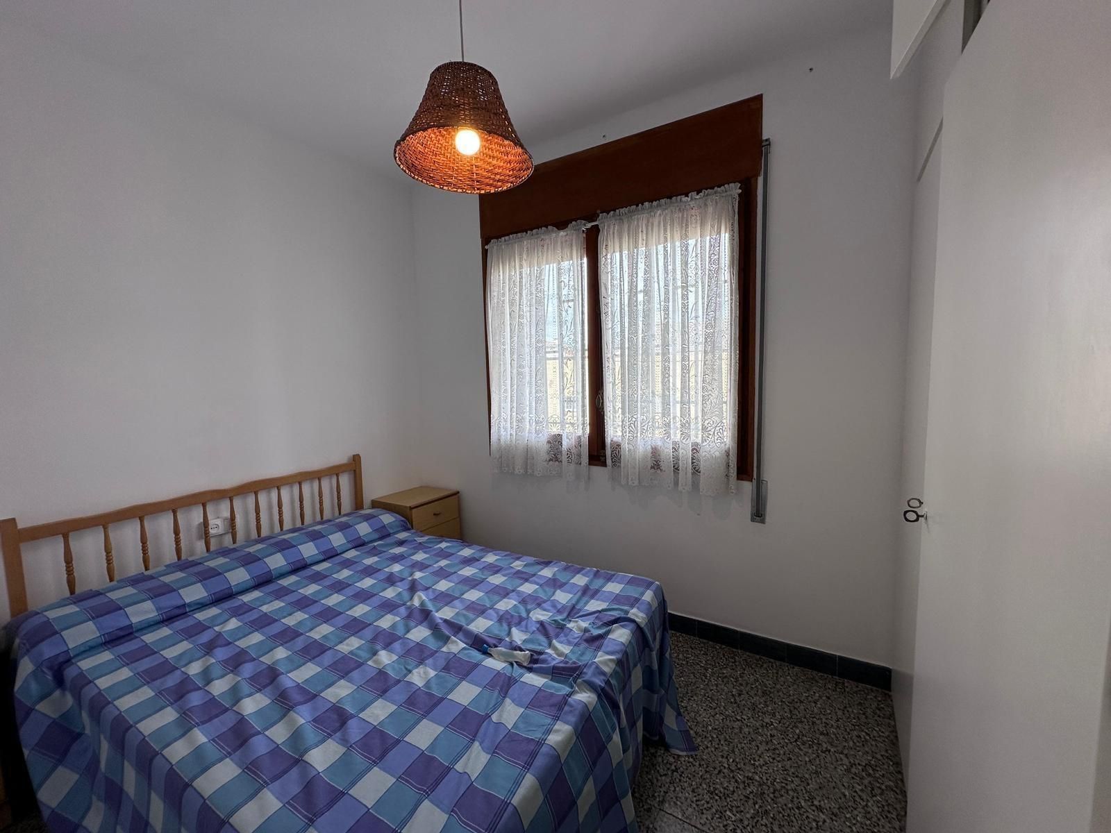 Dormitorio con cama de matrimonio cubierta con sábanas de cuadros azules, ventana con cortinas de encaje y lámpara tejida.