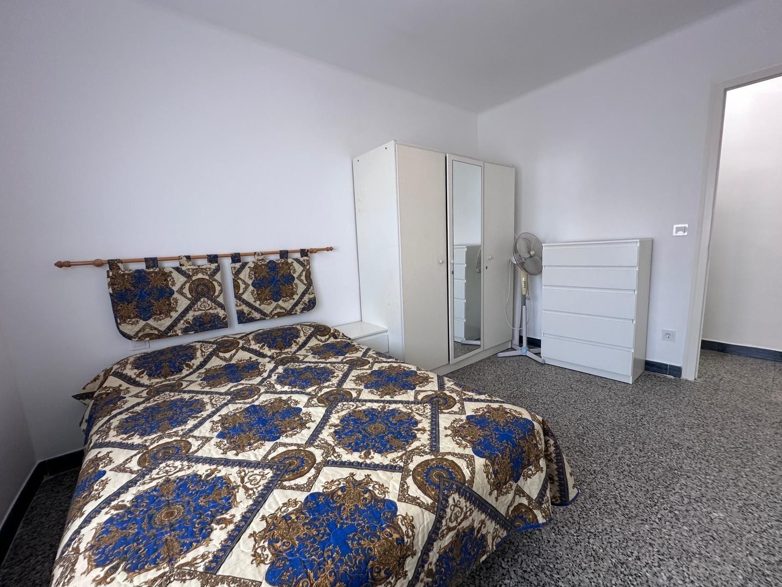 Dormitorio con cama, armario, cómoda y ventilador. Paredes blancas, colcha estampada y suelo moteado oscuro.