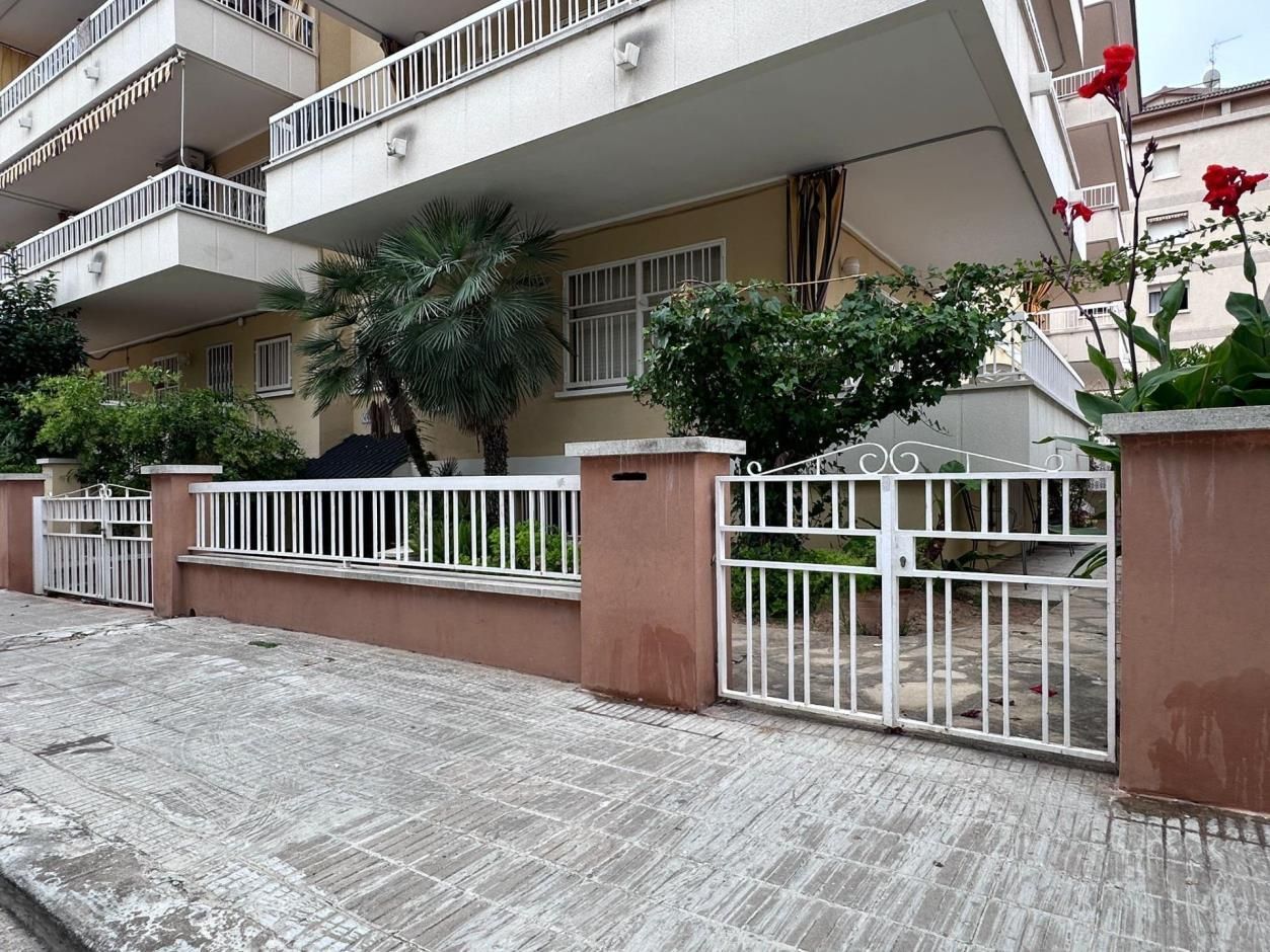 Vista exterior de un edificio de color claro, con balcones, cerca blanca y un portón al frente, con vegetación.