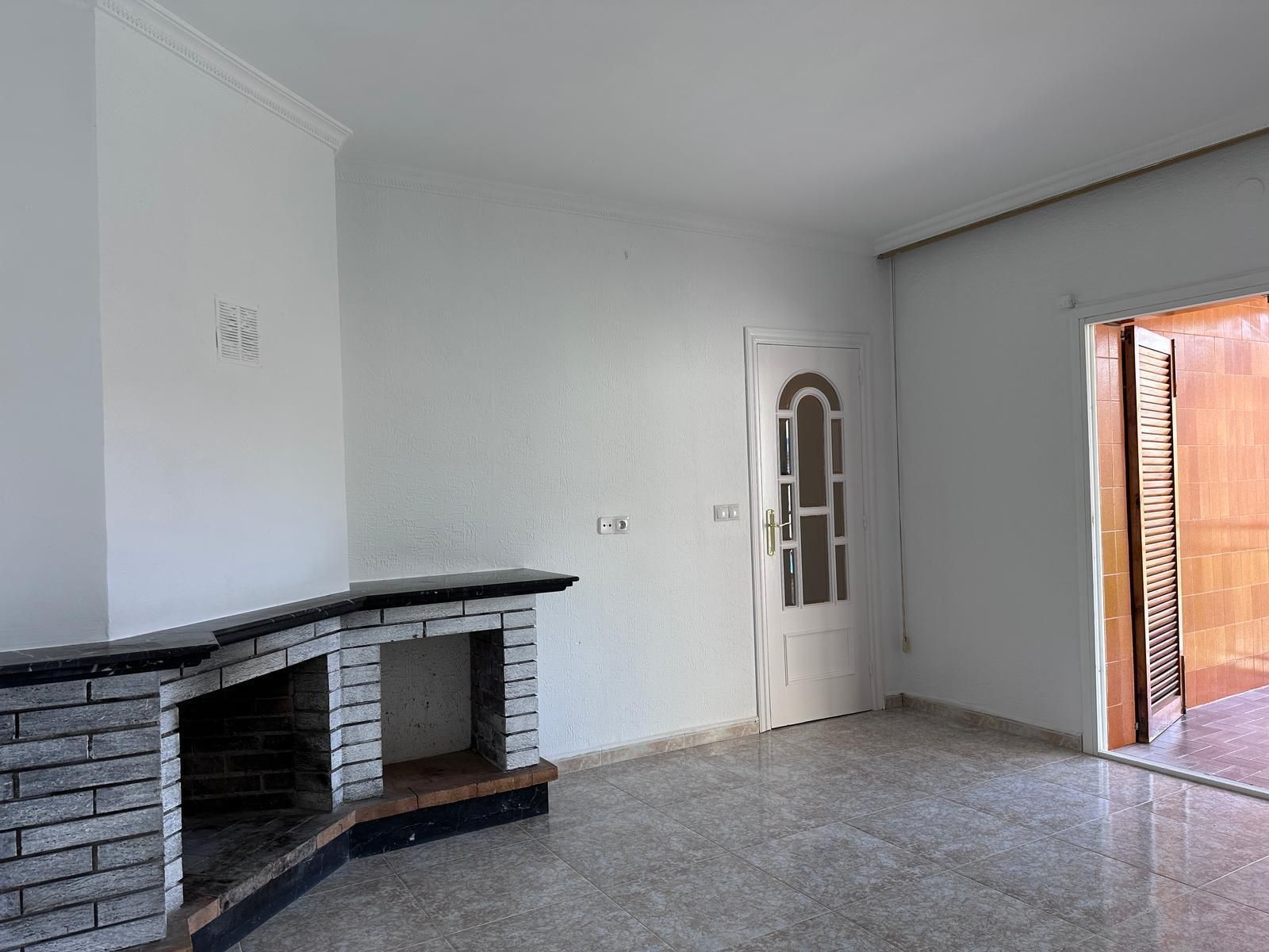 Habitación vacía con chimenea, puerta y entrada abierta, iluminada por luz natural.