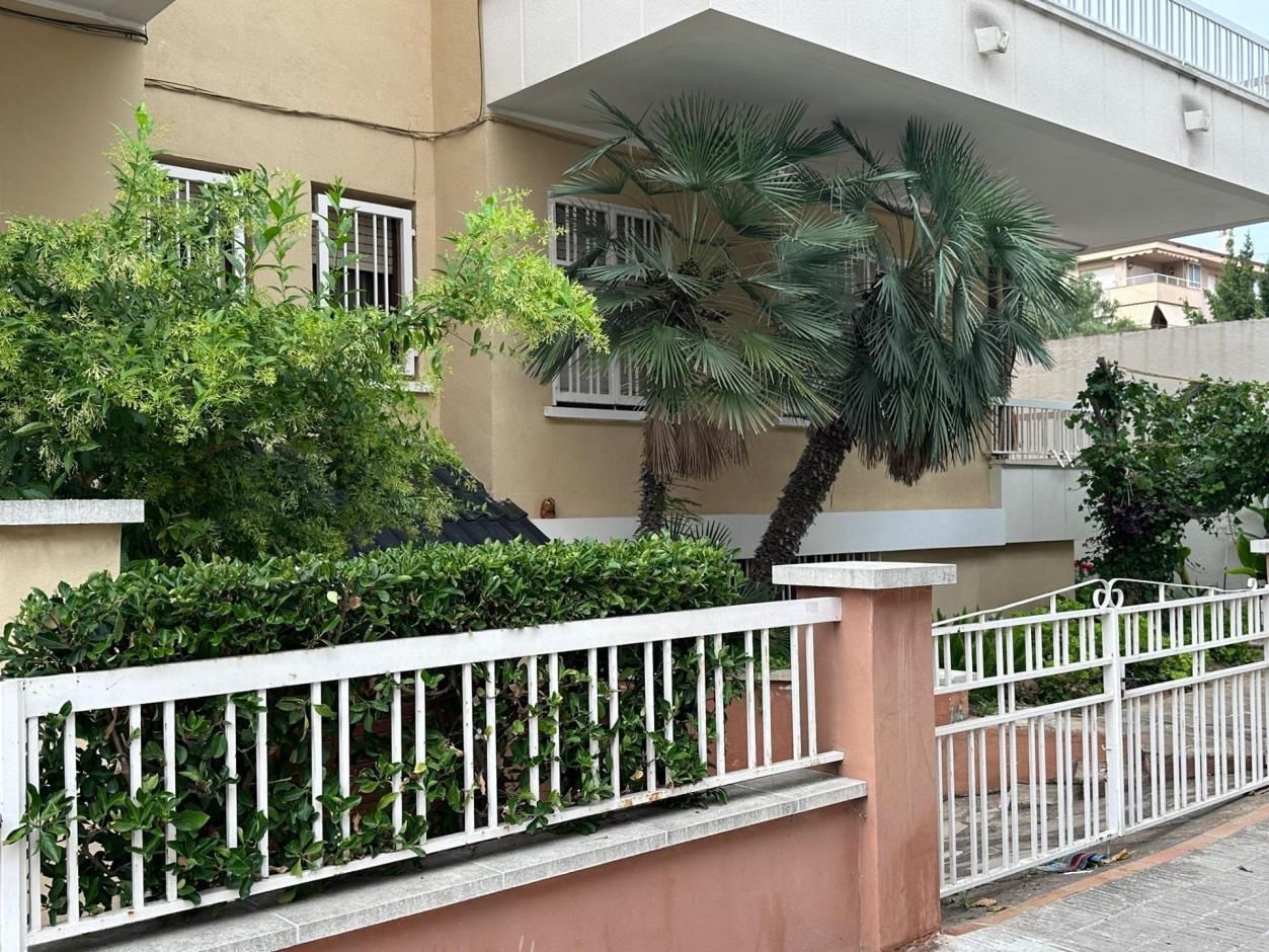Un edificio de dos pisos con molduras blancas, detrás de una cerca blanca y una exuberante vegetación.