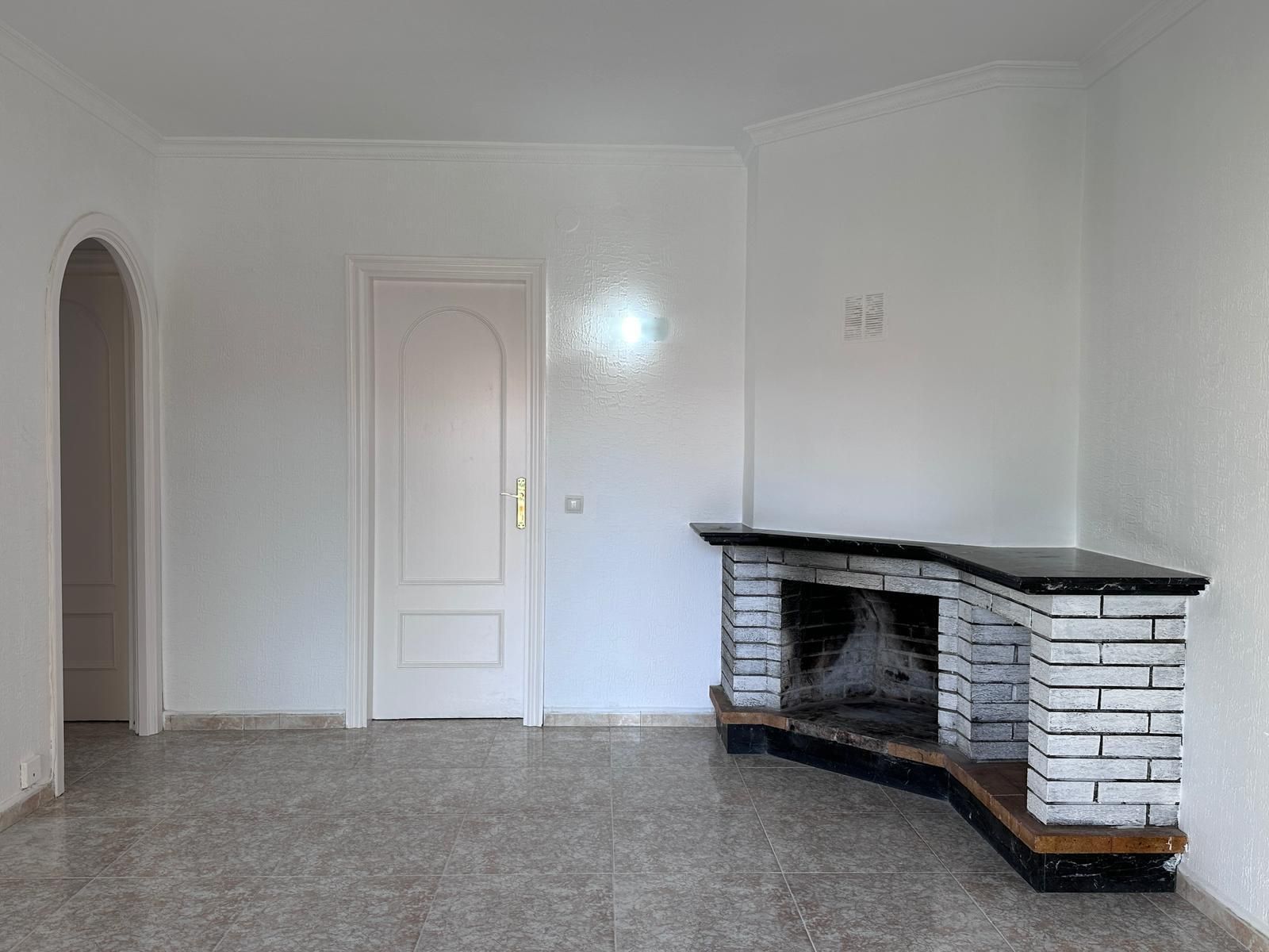 Una habitación de paredes blancas con una chimenea de ladrillo, una puerta y una entrada arqueada, todo sobre un piso de baldosas.