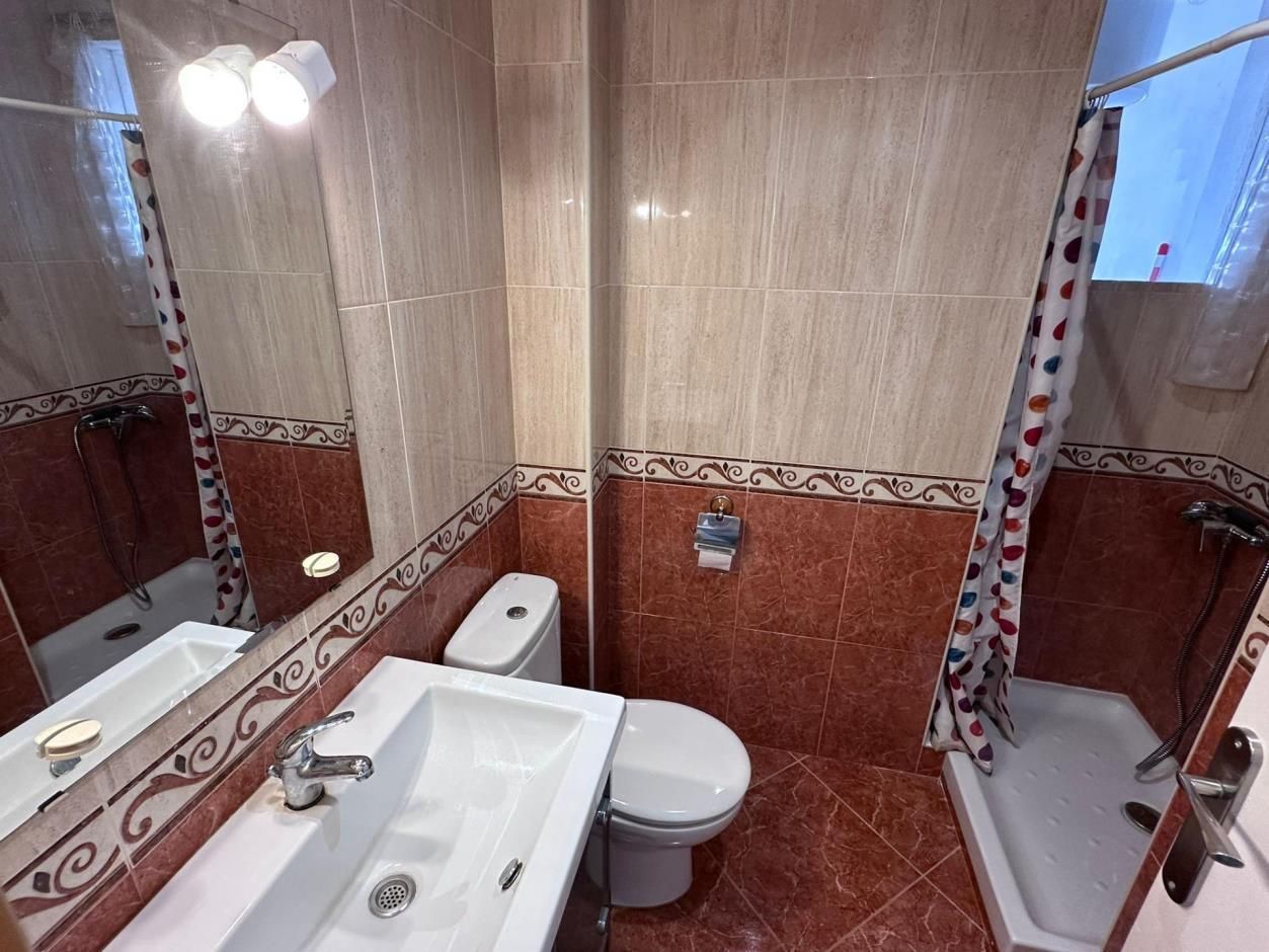 Pequeño baño con paredes de azulejos de color rojo y tostado, inodoro, lavabo, ducha y espejo.