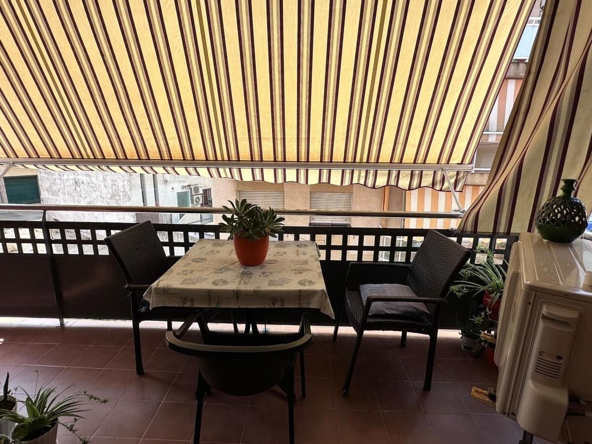 Un pequeño balcón con mesa, sillas y una planta en maceta, bajo un toldo de rayas.