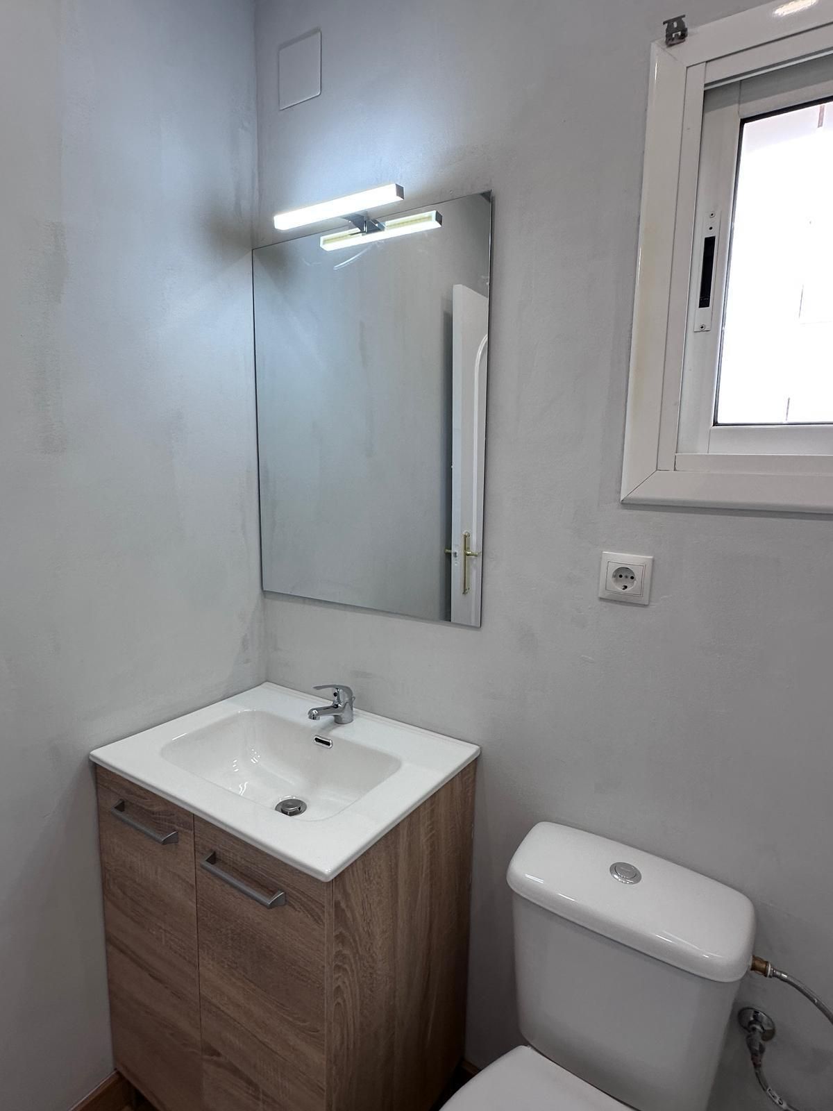 Baño pequeño con tocador de madera, inodoro, espejo y ventana. Paredes grises y grifería blanca.