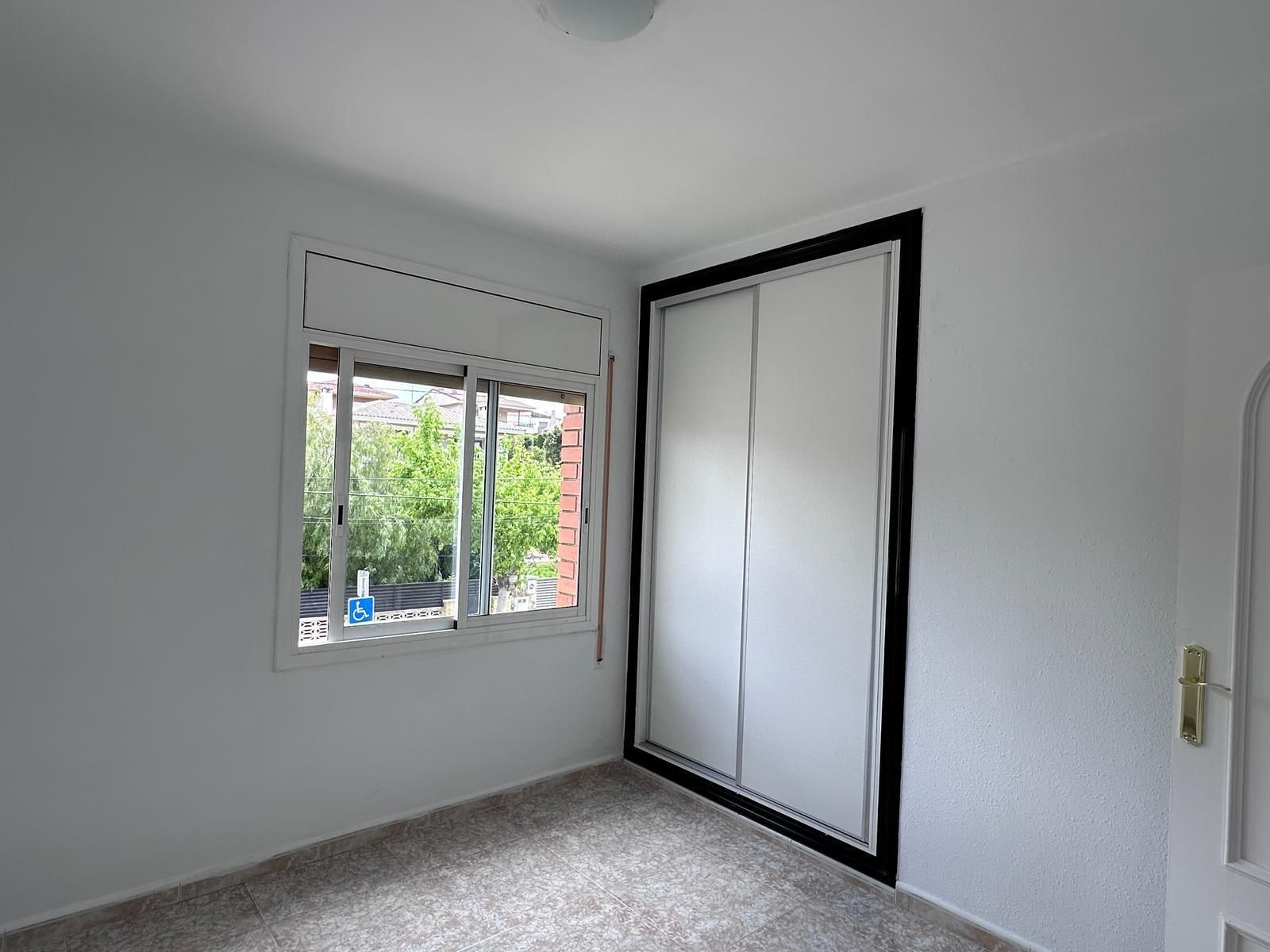 Habitación vacía con ventana y armario. Paredes blancas, suelo alfombrado. Se ve follaje verde por la ventana.