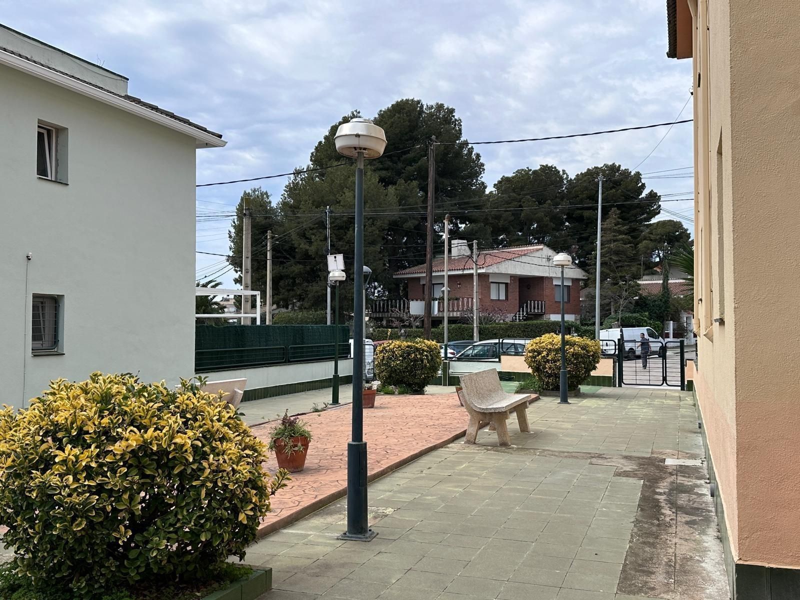Un patio con un banco, arbustos y una farola. Edificios y árboles al fondo bajo un cielo nublado.