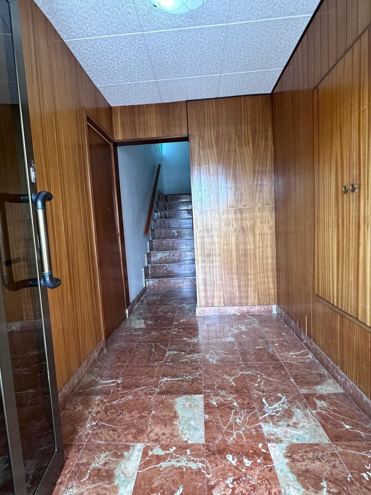 Pasillo estrecho con suelo de baldosas rojas, paredes revestidas de madera y escaleras que suben. Hay una puerta de cristal a la izquierda.