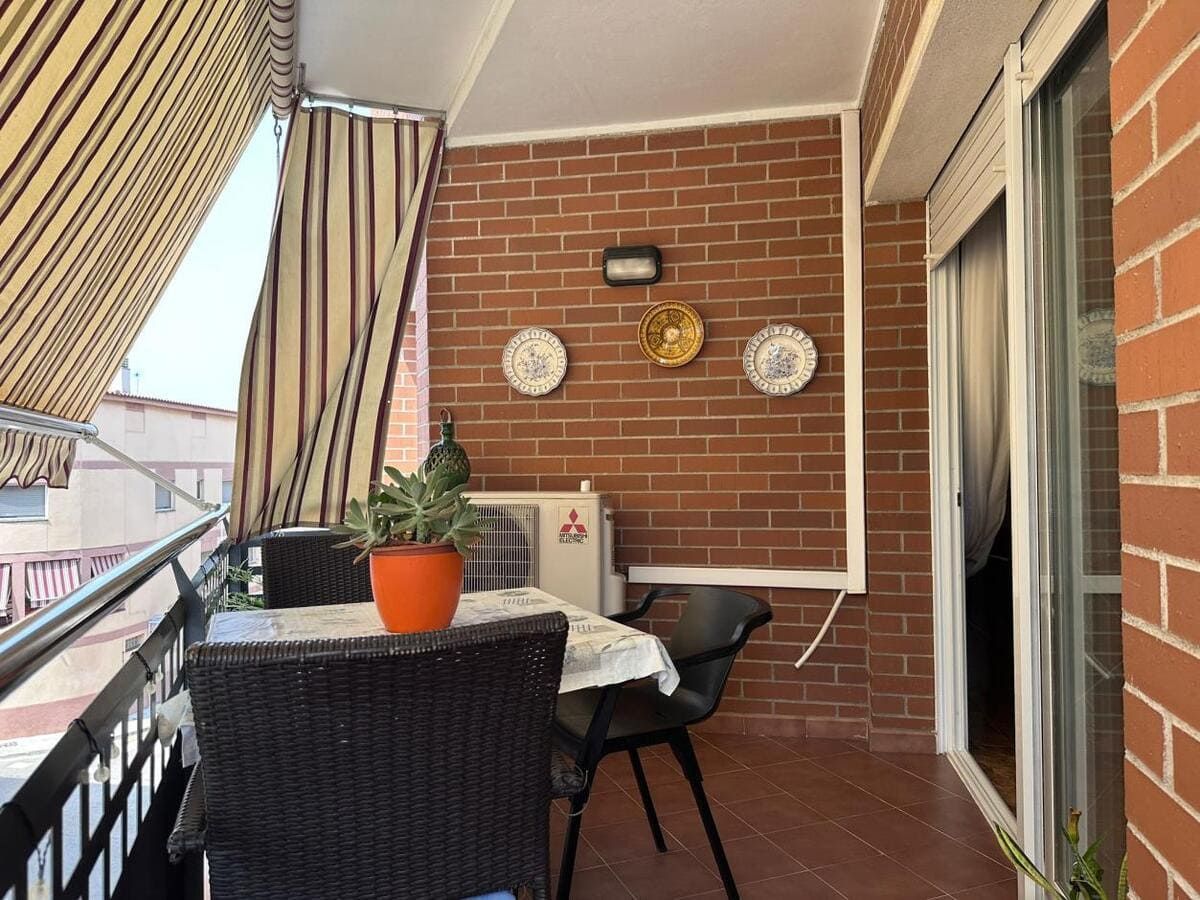 Balcón con mesa, sillas y maceta. Pared de ladrillo con platos decorativos y aire acondicionado.