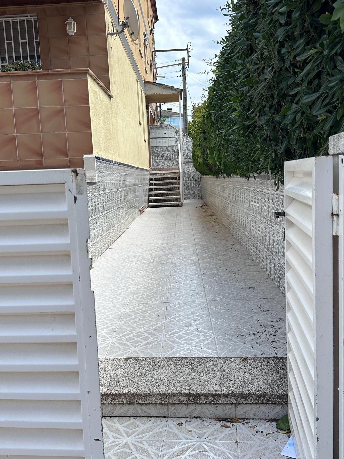 Una puerta blanca abierta conduce a un largo camino de baldosas entre un edificio y un seto verde; escalones al final.