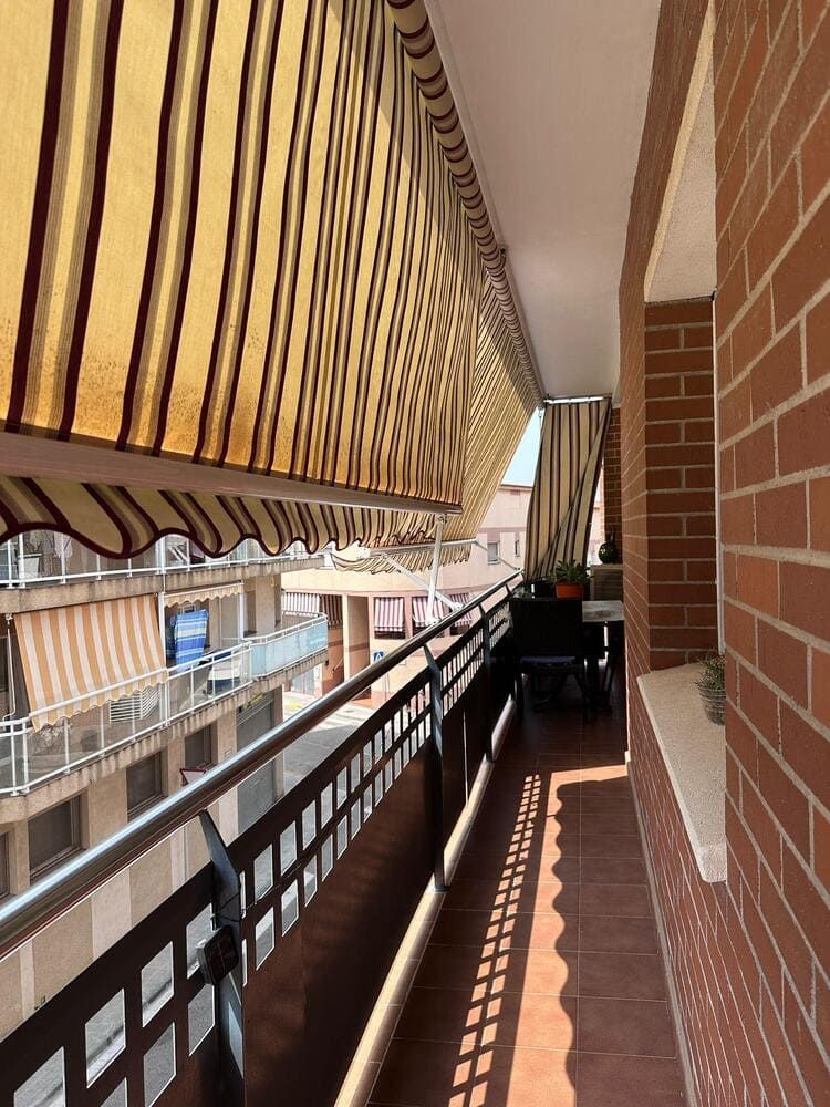 Balcón con toldo a rayas. Pared de ladrillo rojo a la derecha, barandilla metálica y vistas a los edificios.