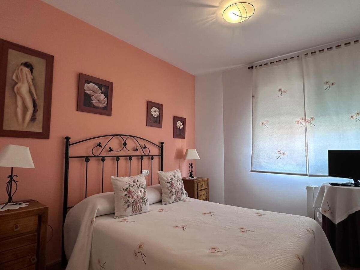 Dormitorio con paredes color durazno, marco de cama de hierro forjado, obras de arte florales, mesitas de noche y televisor.