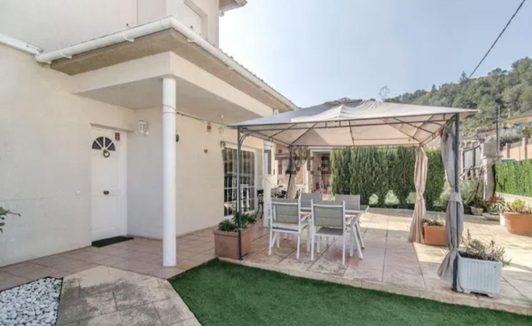 Exterior de casa blanca con patio y cenador, mesa con sillas, seto verde y plantas en macetas.