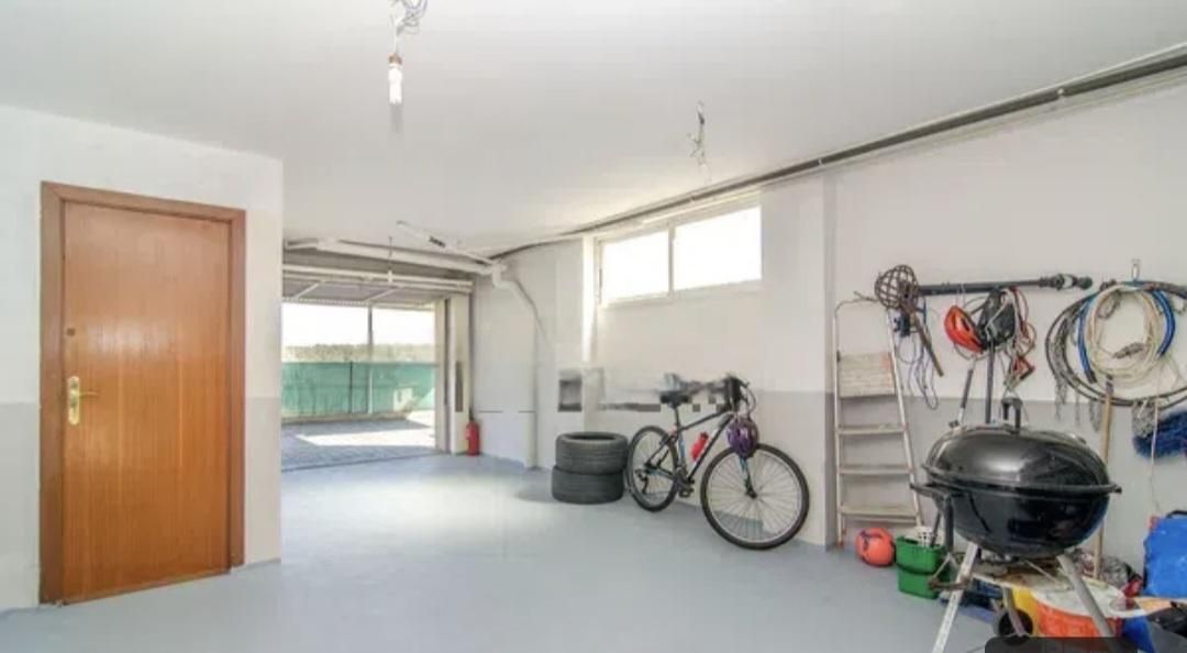 Interior de garaje con bicicleta, parrilla y almacenamiento, piso azul claro, puerta a la izquierda, puerta de garaje abierta al fondo.