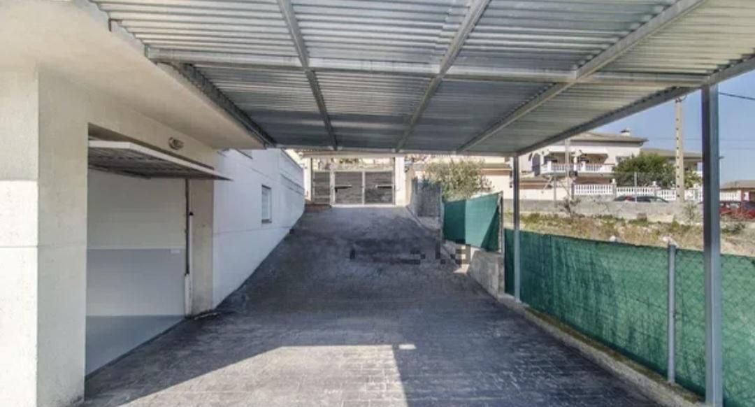 Un camino de entrada bajo un toldo metálico conduce a una entrada cerrada. Edificio blanco a la izquierda, valla verde a la derecha.