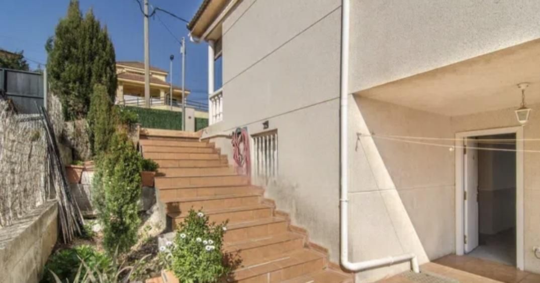 Vista exterior de una casa de dos pisos con escaleras de ladrillo que conducen a una puerta y un jardín.