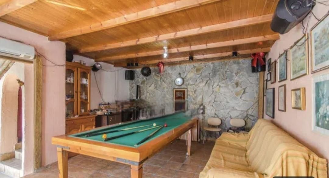 Sala de billar con mesa de billar, pared de piedra, sofá y cuadros enmarcados; techo de madera y paredes rosas.