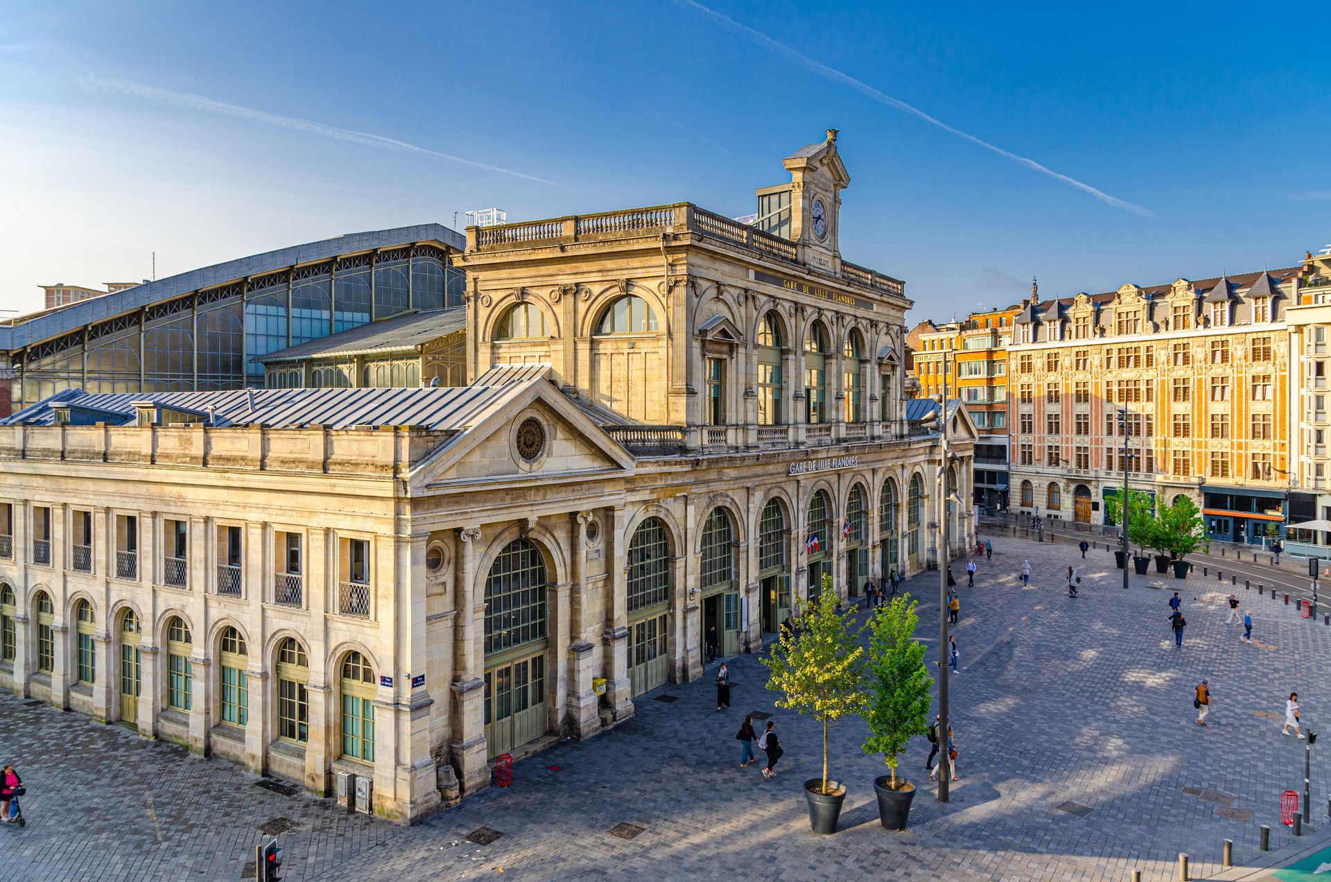 Bâtiment historique en pierre avec des fenêtres cintrées jouxtant une place moderne, sous un ciel bleu clair.