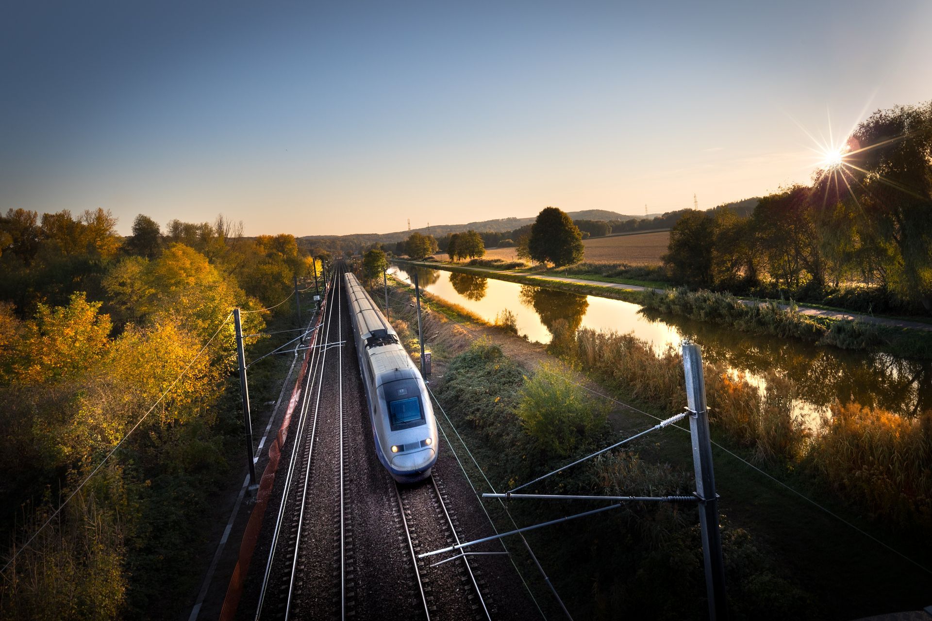 Train à grande vitesse traversant un pont au bord d'une rivière au coucher du soleil, avec des arbres d'automne et des lignes électriques.