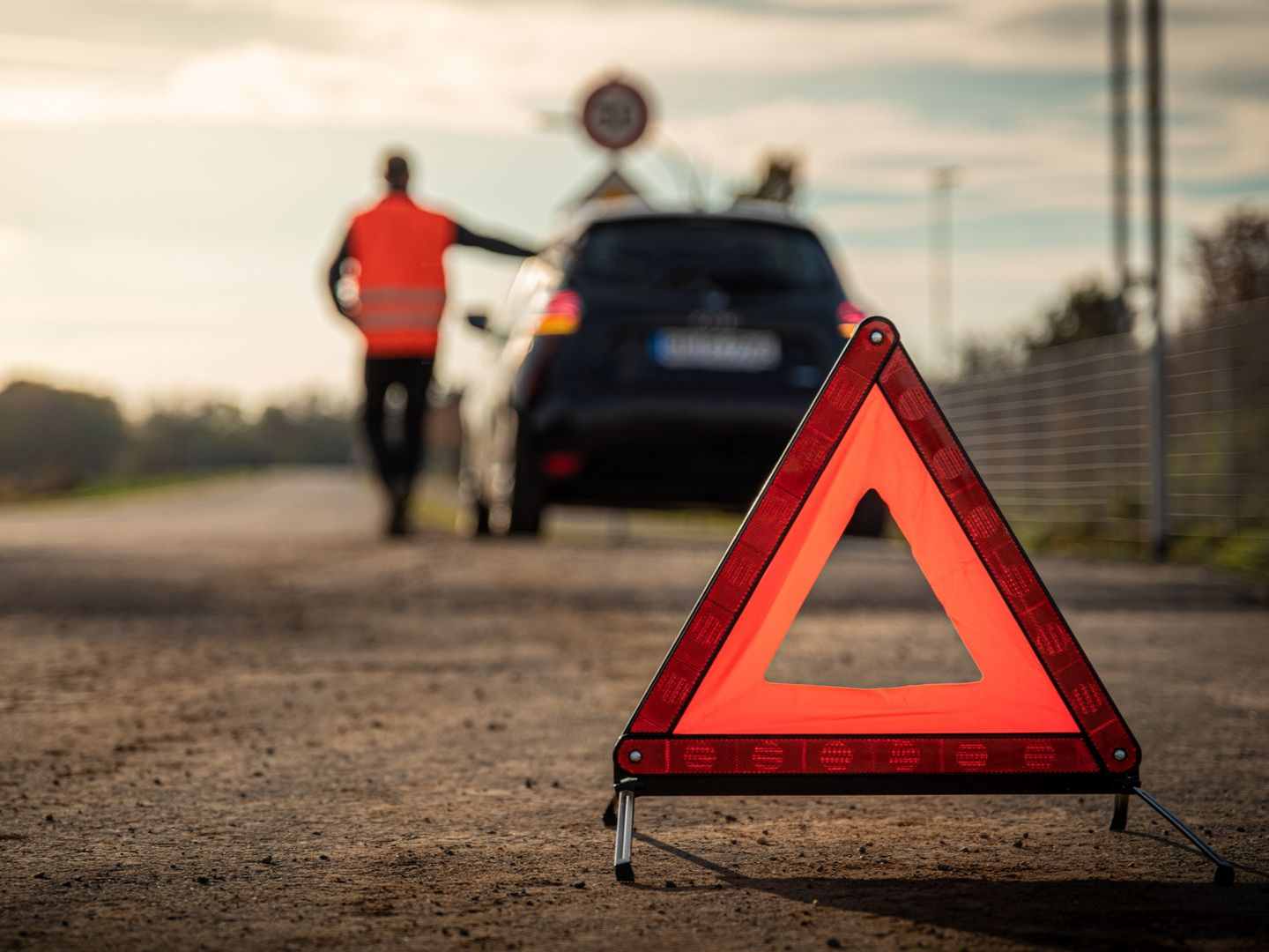 Triangle de signalisation rouge à côté d'une voiture en panne et d'une personne sur le bord de la route.