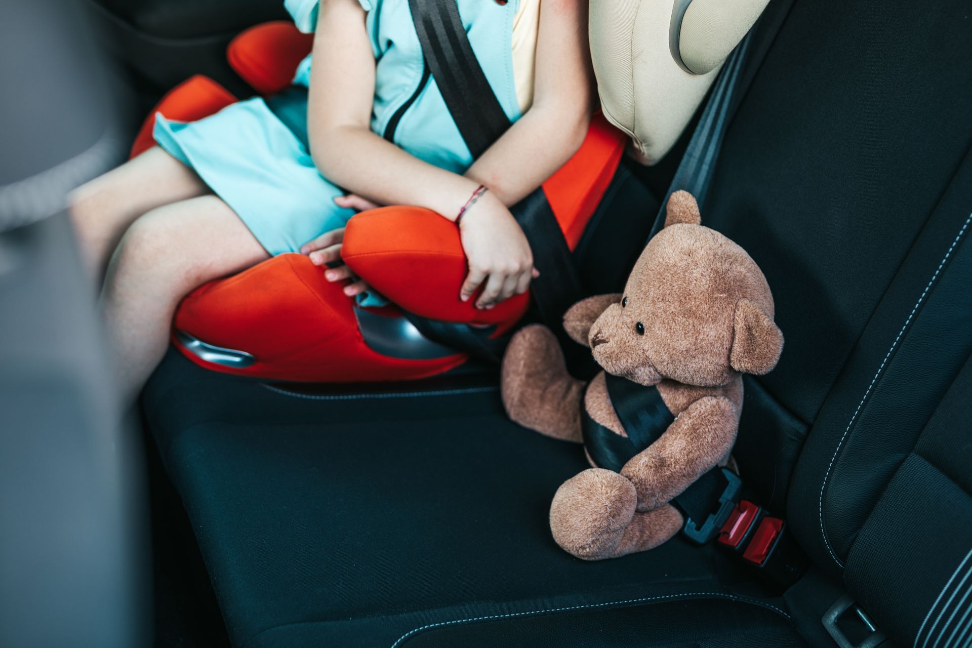 Un enfant dans un siège auto rouge avec un ours en peluche à côté de lui.