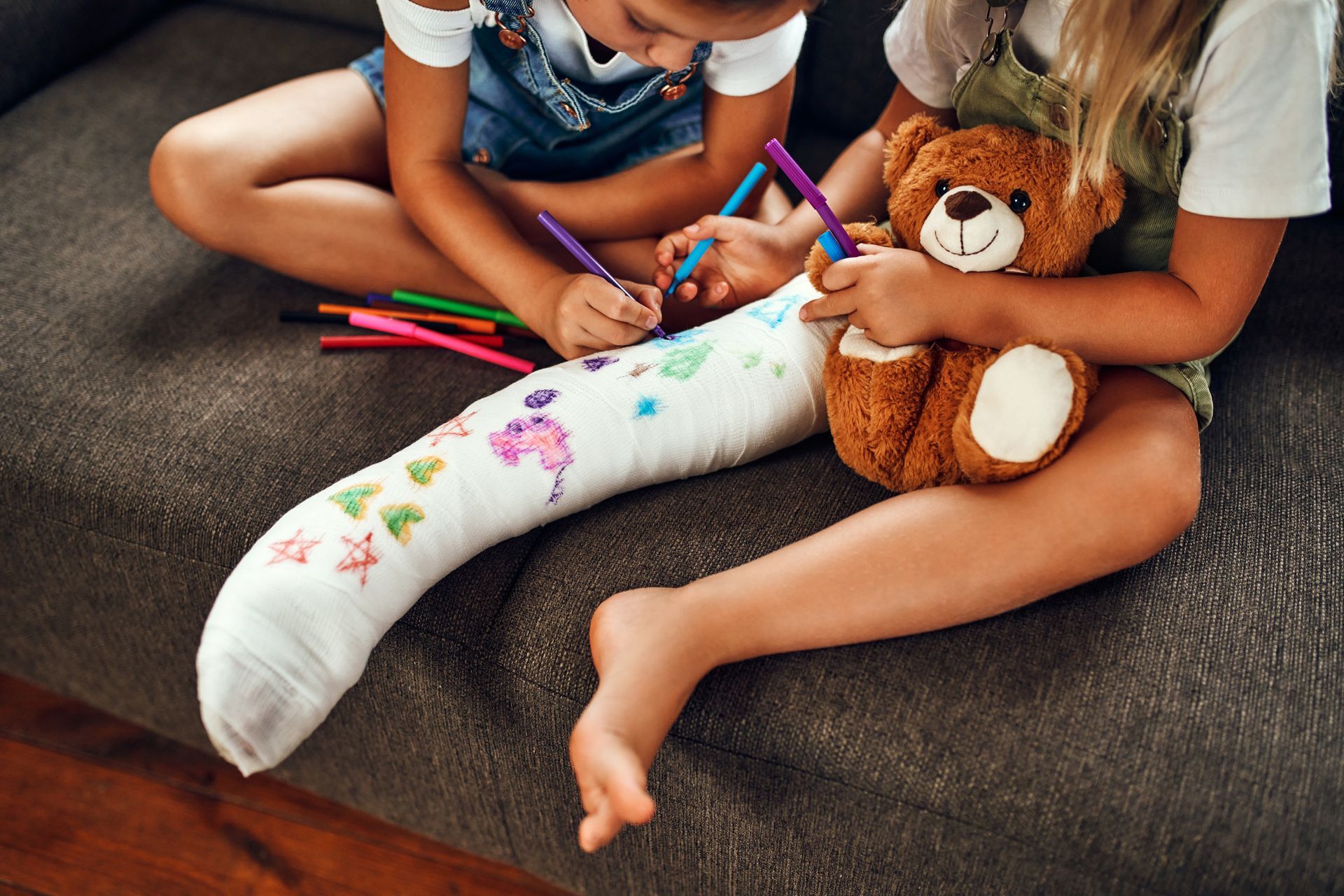 Deux enfants colorient un plâtre tout en tenant un ours en peluche sur un tapis gris.