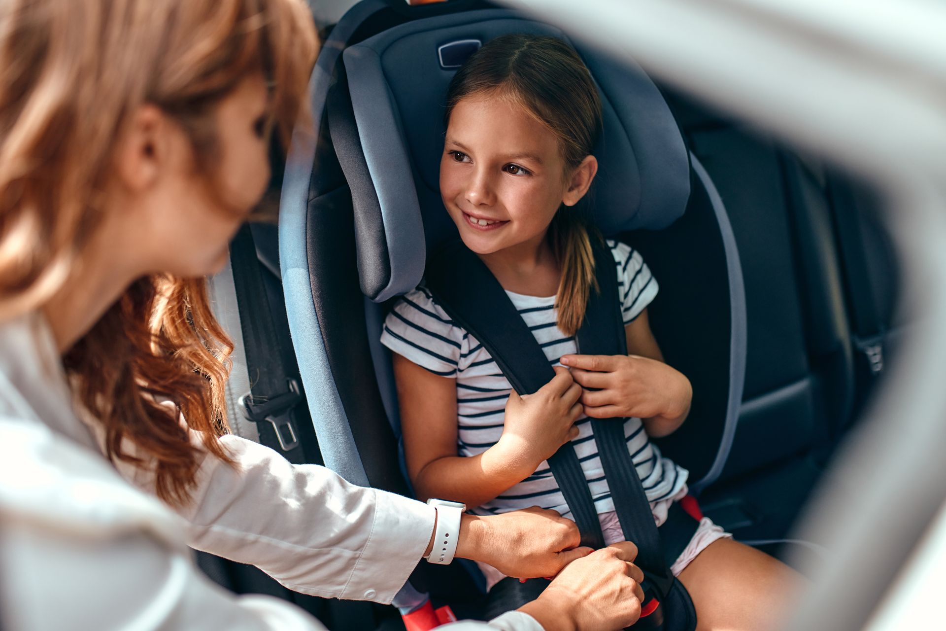 Un adulte attache la boucle du siège auto d'un enfant ; l'enfant sourit dans un siège auto gris.