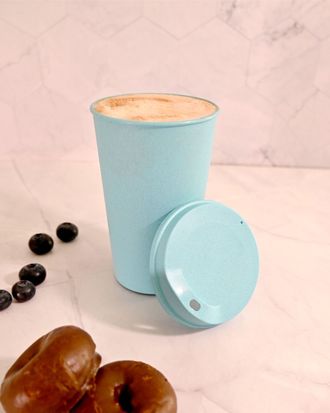Taza azul claro con tapa, junto a granos de café y dos dátiles sobre una superficie de mármol.