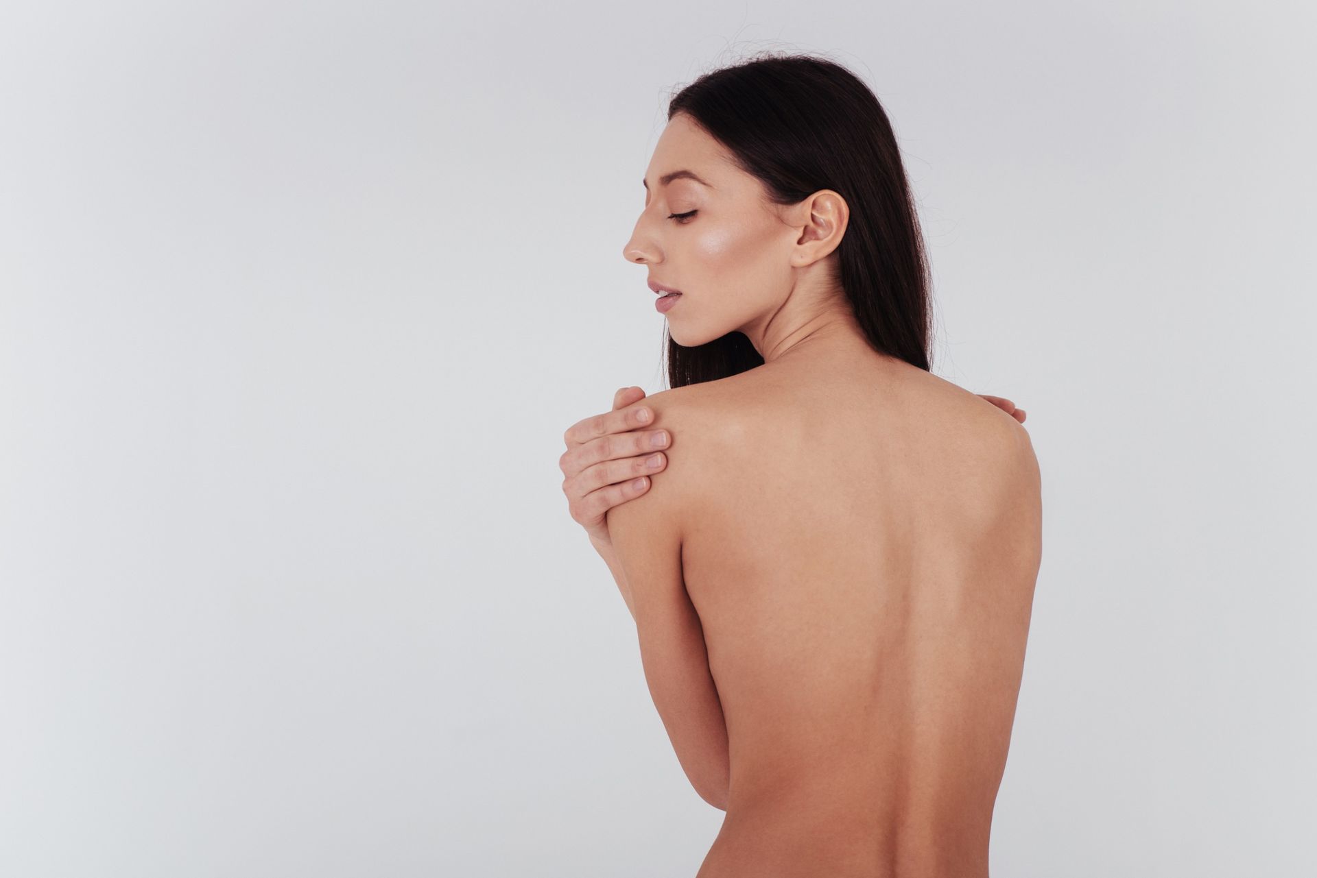 Mujer con cabello largo y oscuro, de espaldas a la cámara, con la espalda descubierta y las manos cruzadas, fondo blanco.