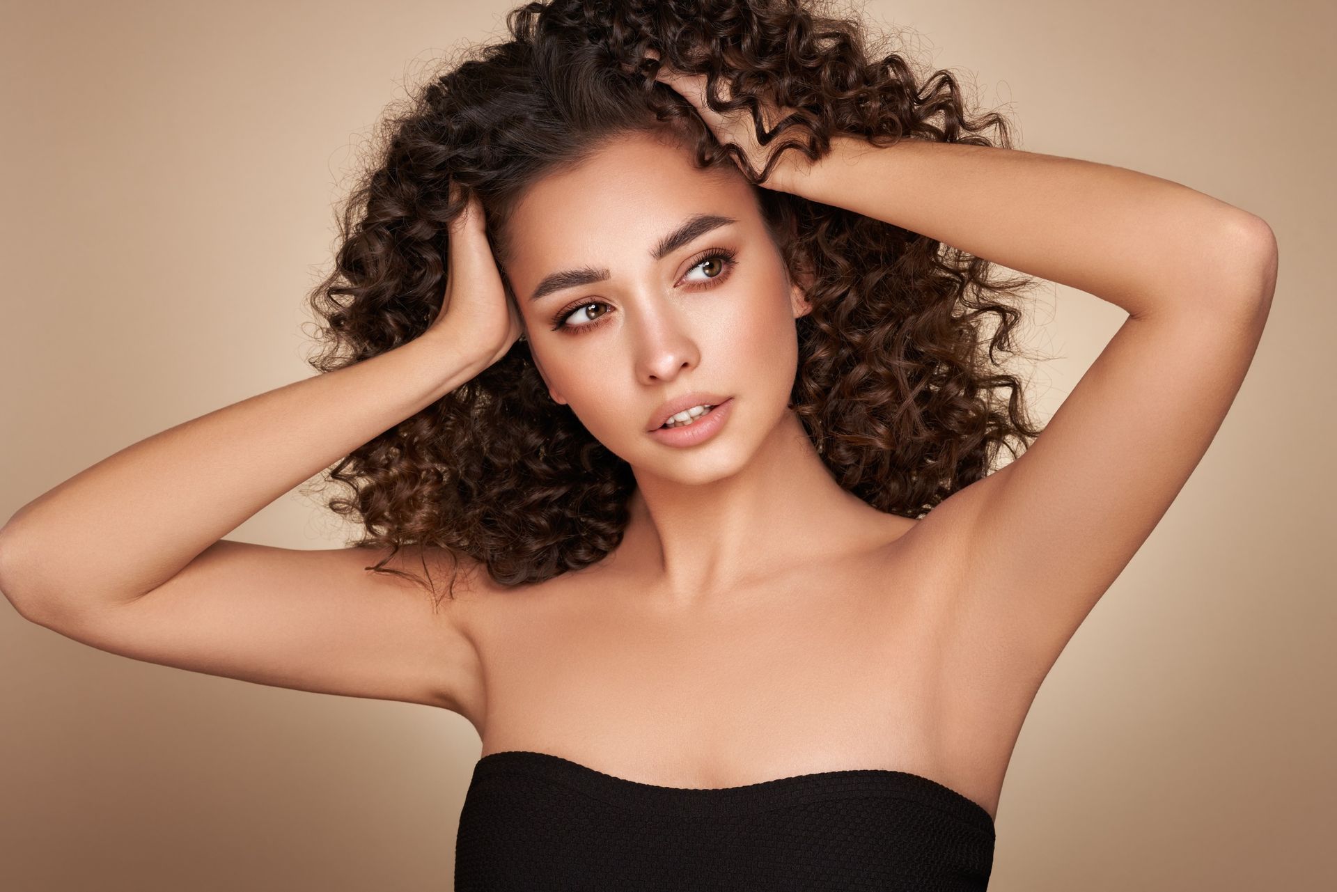 Mujer de cabello castaño rizado con top negro sin tirantes, con las manos en el pelo y la mirada perdida. Fondo beige.