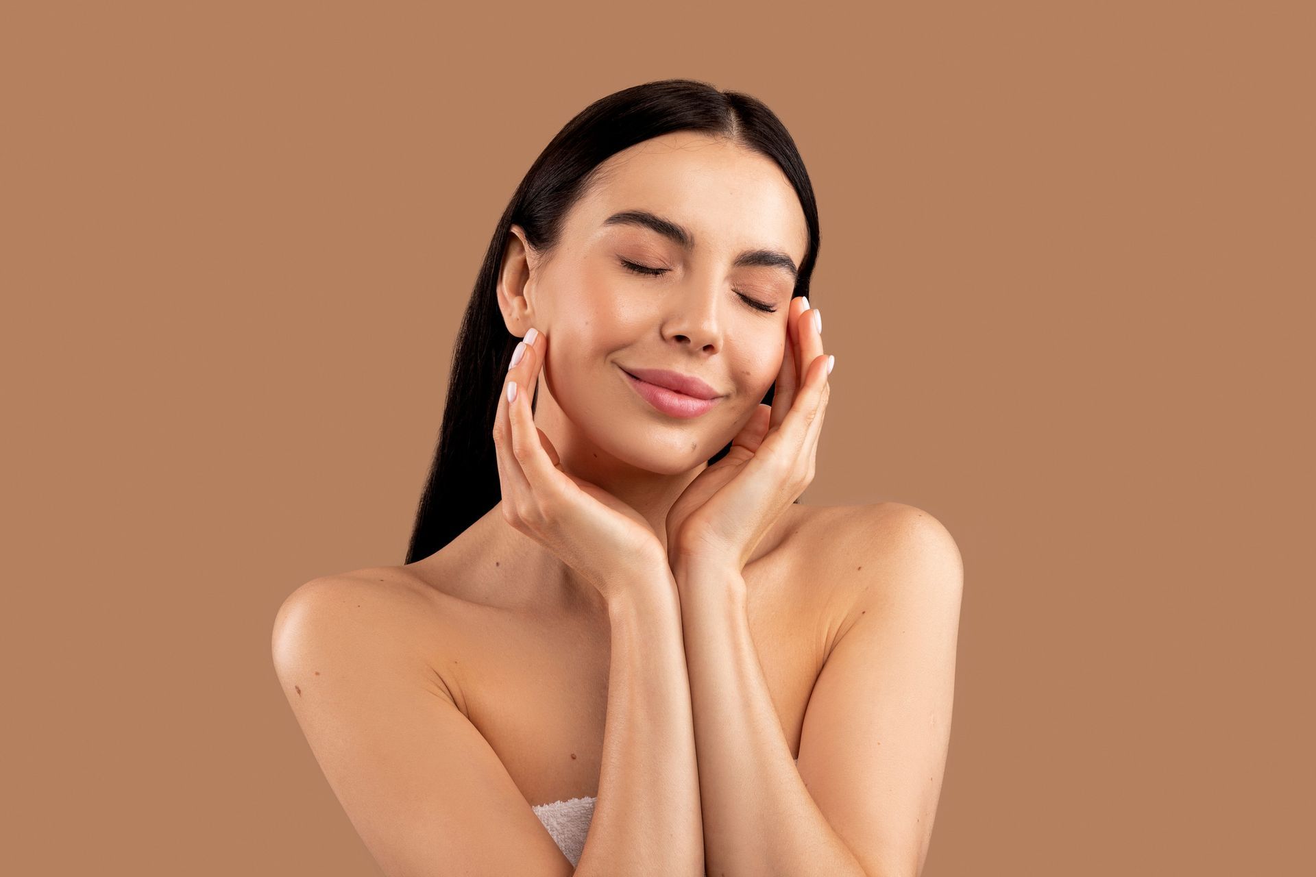 Mujer de cabello y ojos oscuros, sonriendo, tocándose la cara con ambas manos sobre un fondo neutro.
